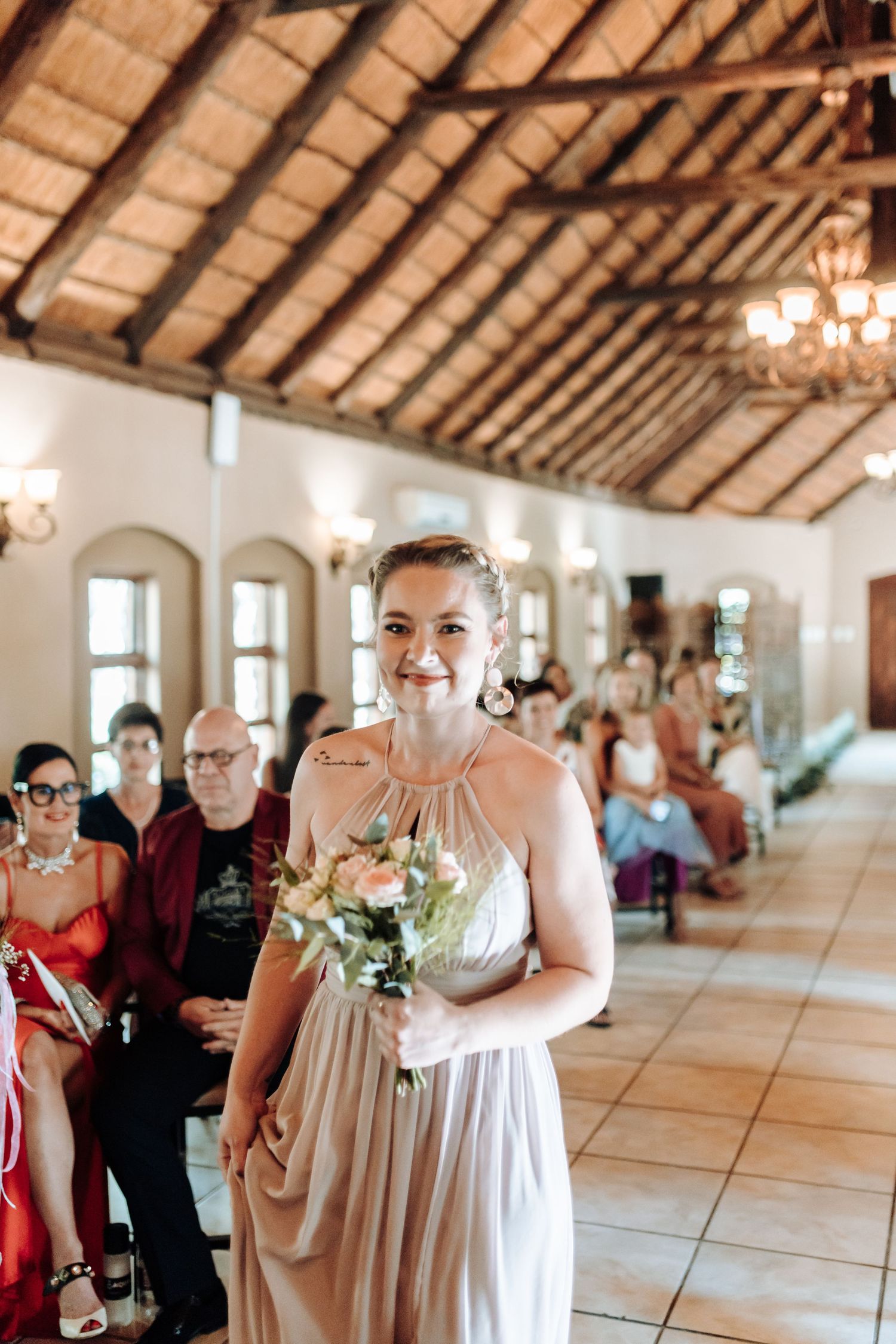A bridesmaid holds a bouquet while standing in a rustic venue with vaulted ceilings and chandeliers.
