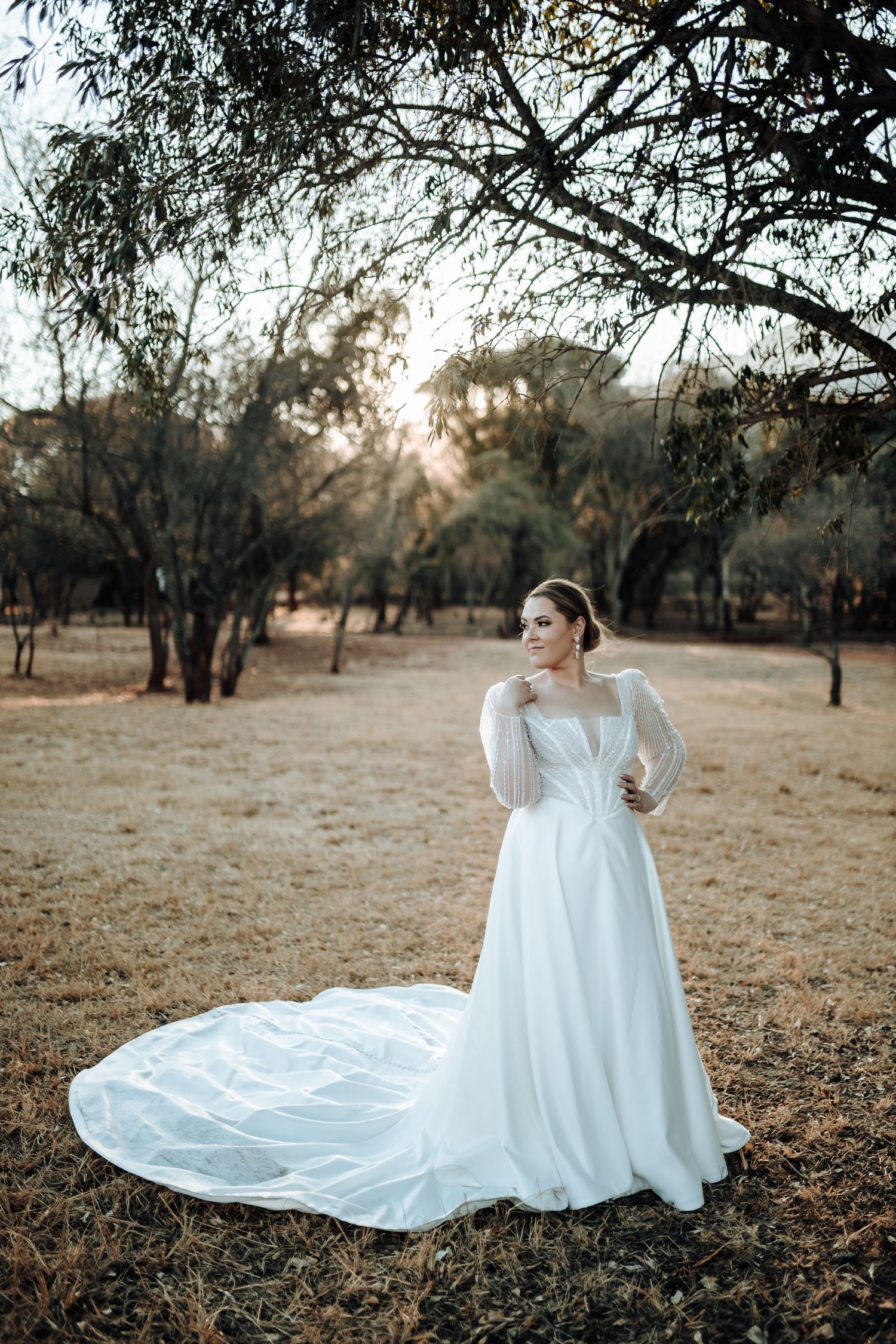 Elegant bride in long sleeve white gown with flowing train poses in autumn field at sunset.