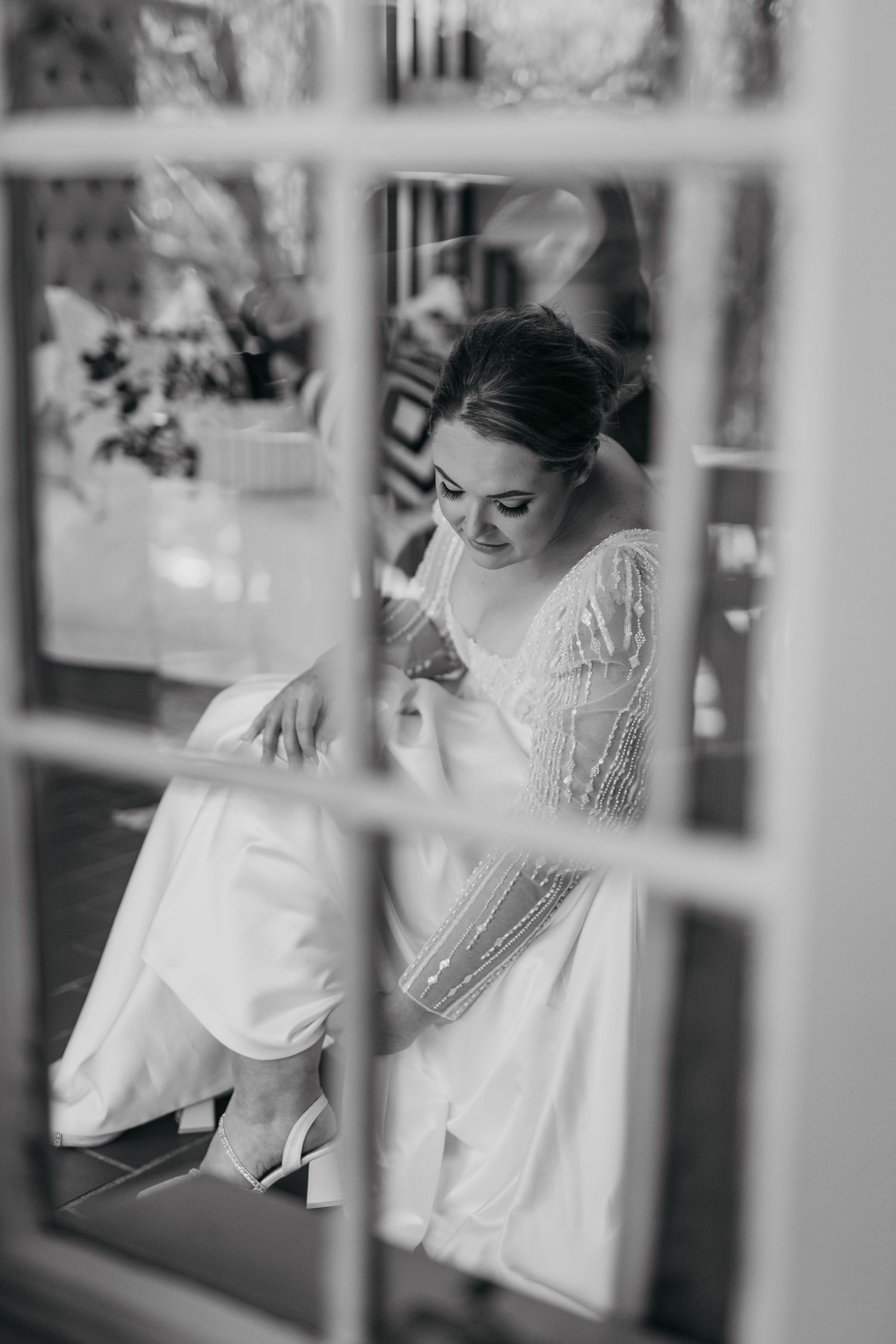 Black and white photo of bride in beaded gown viewed through window panes.