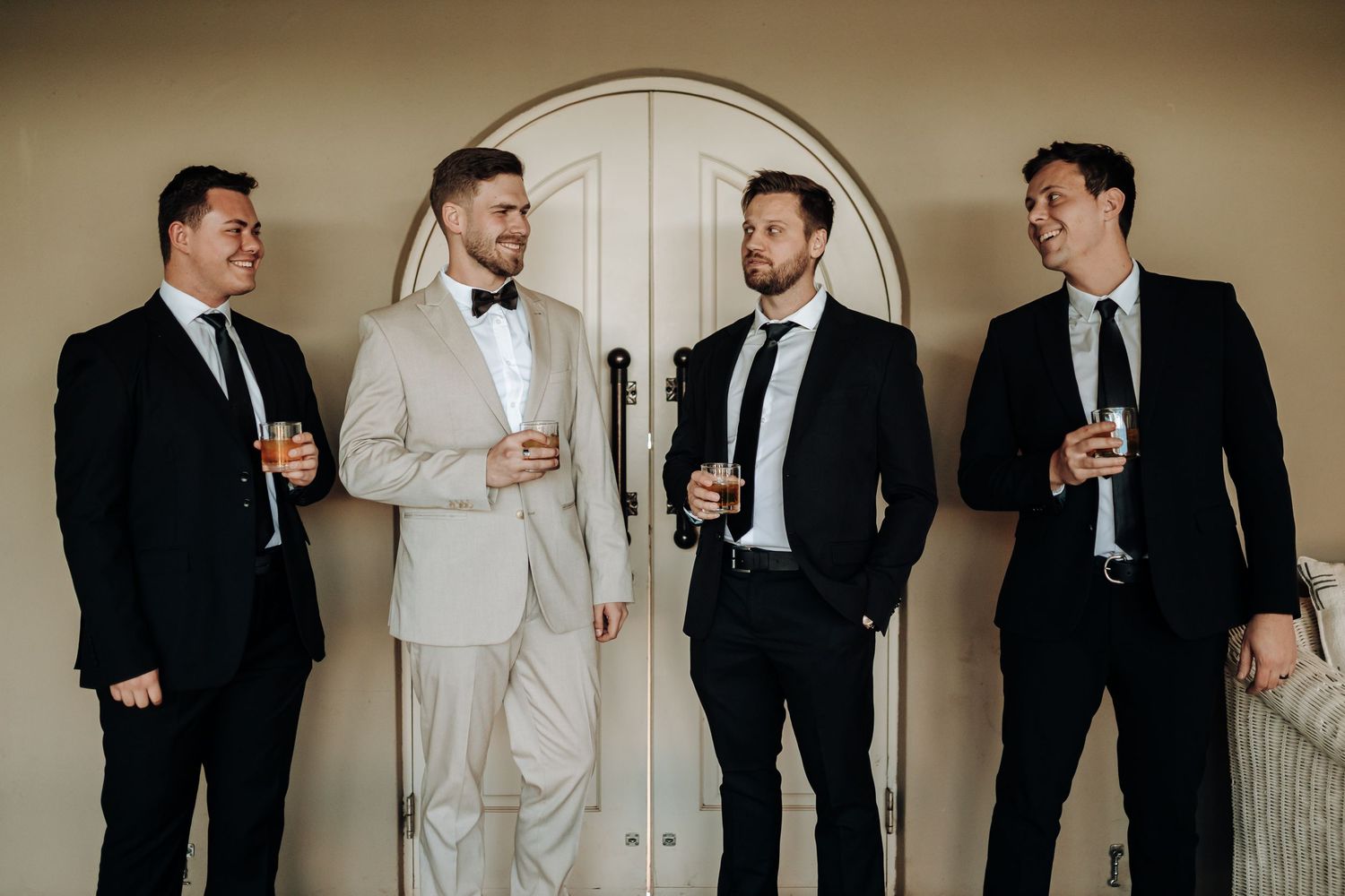 Groomsmen in classic suits gather for drinks beneath an arched doorway at a wedding celebration.