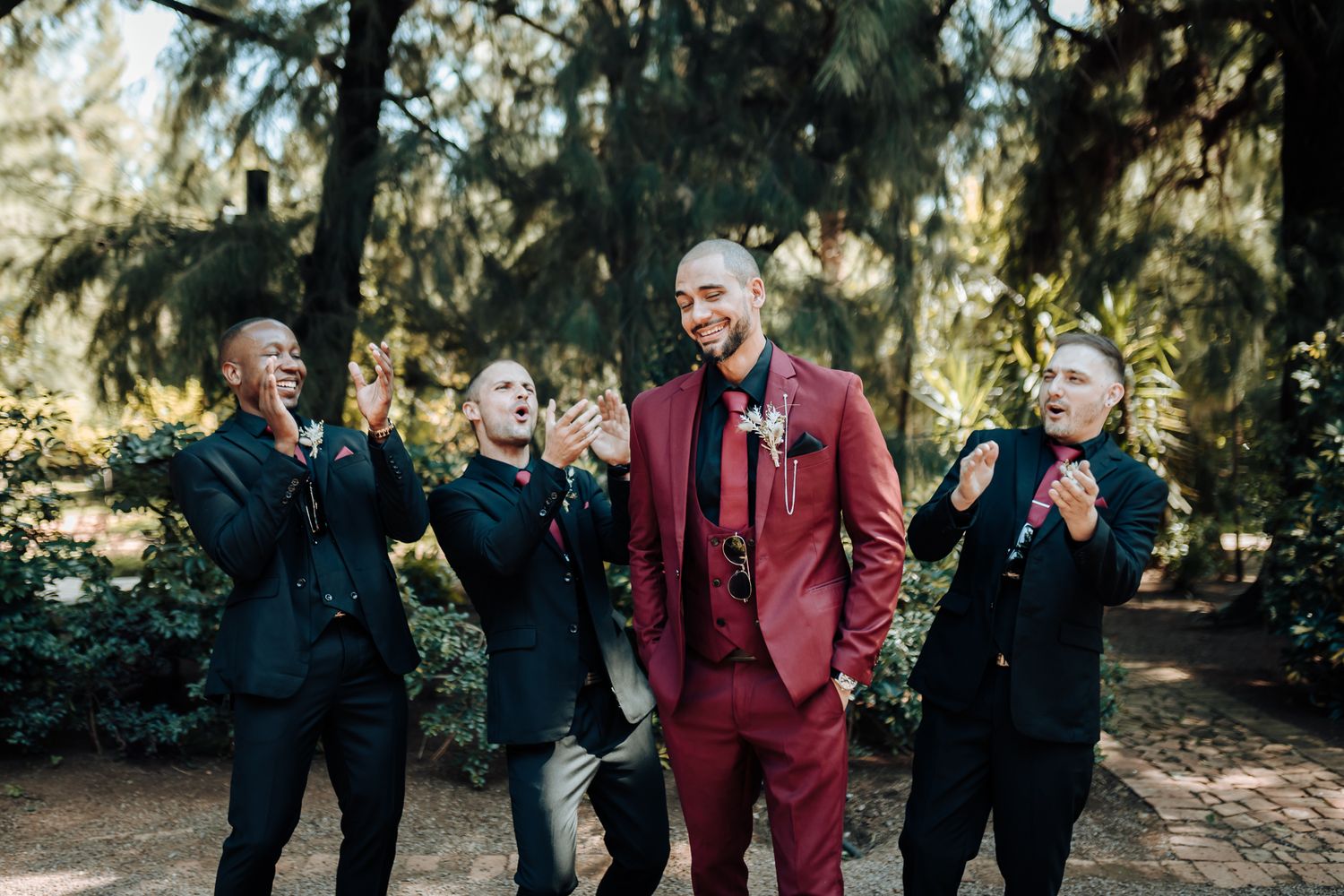 Groomsmen in black suits playfully pose with groom in burgundy suit.