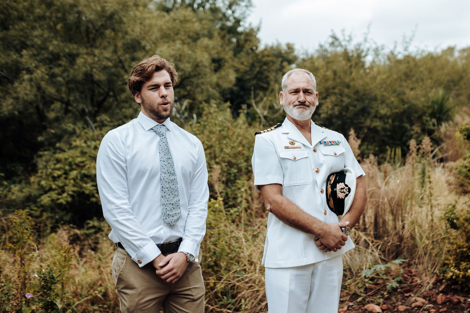 Wedding portrait featuring two people in formal attire standing outdoors among tall grass.