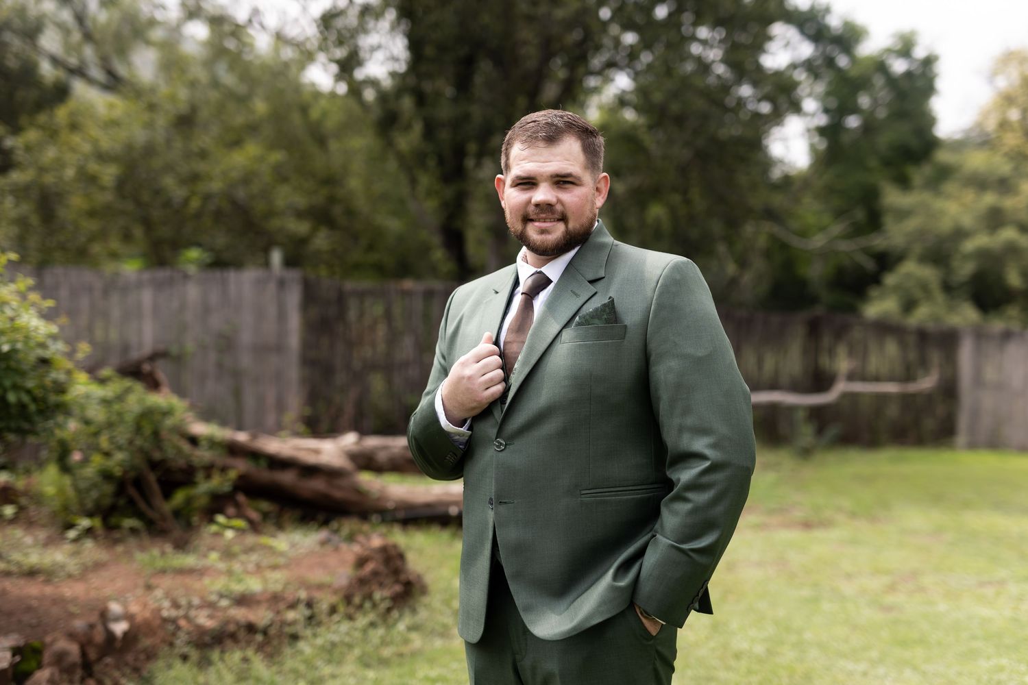 Groom in green suit jacket poses casually in outdoor garden setting.