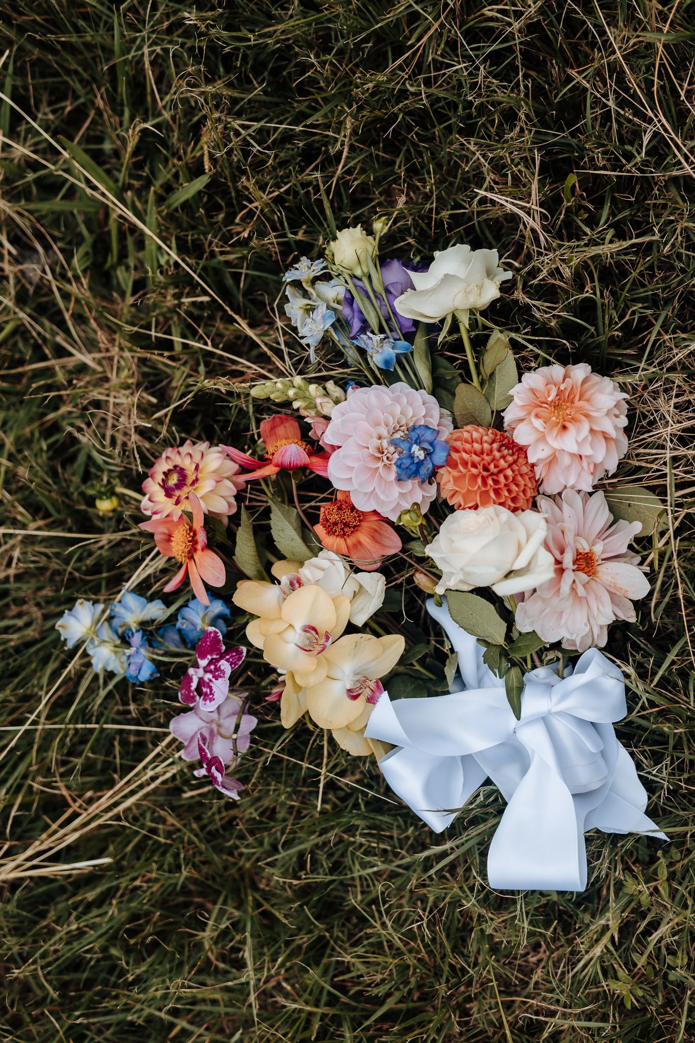 A romantic bridal bouquet with coral, purple and white flowers tied with a white satin bow rests on dried grass.