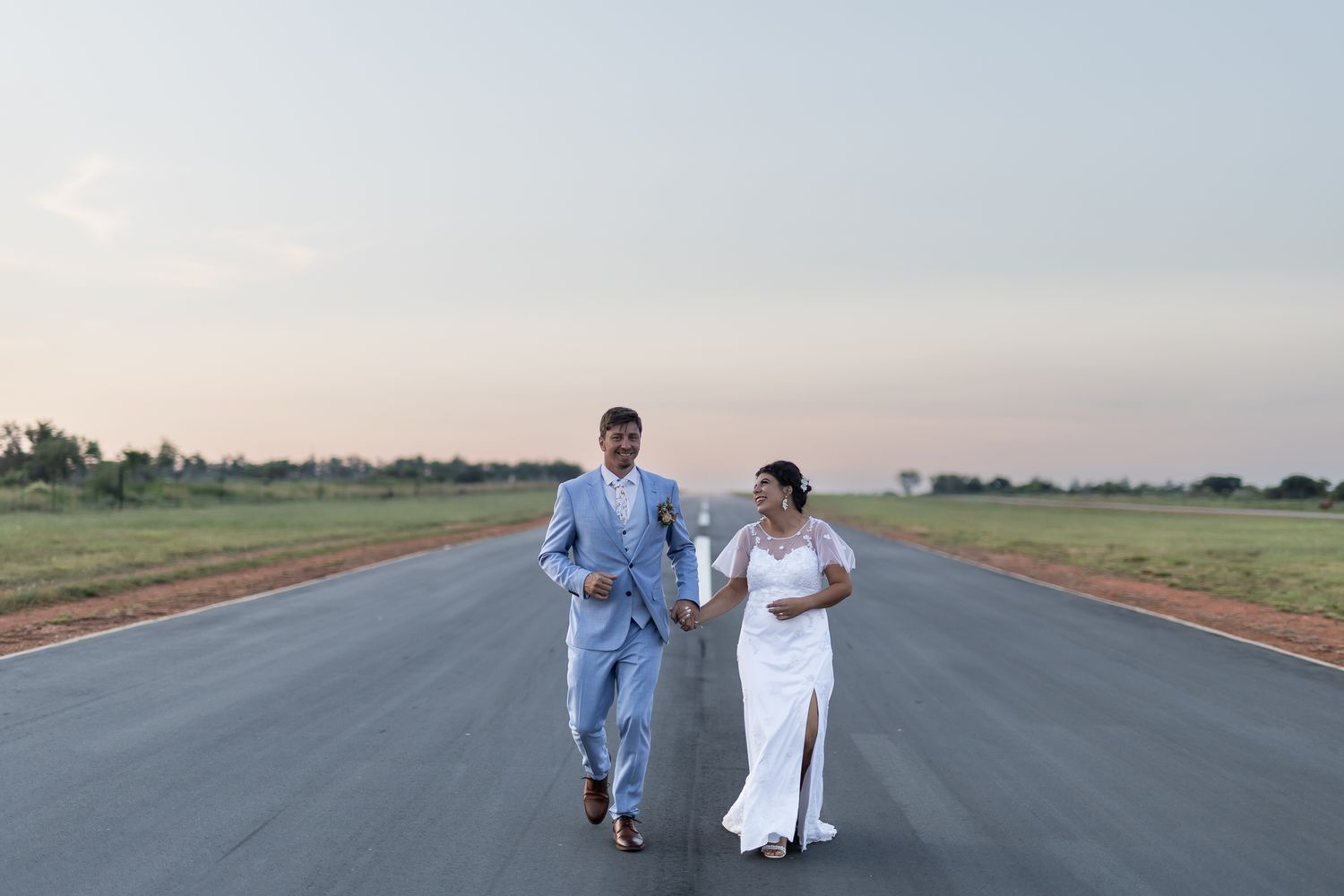 Bride and groom running together on empty runway during sunset.