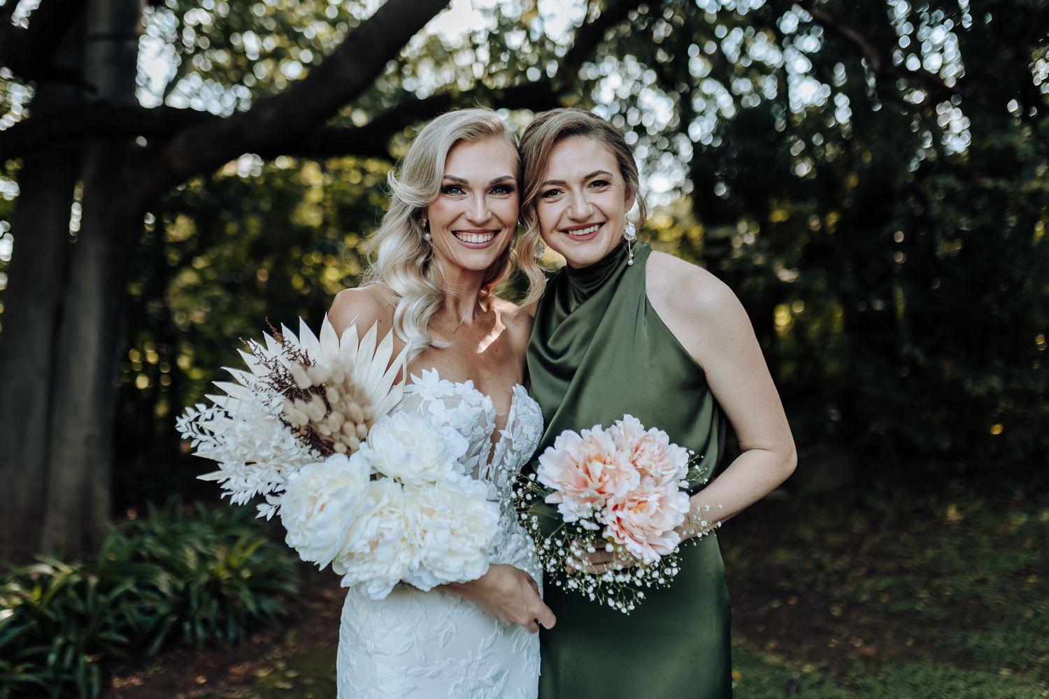 Wedding photo of two people holding bouquets with white and pink flowers against trees.