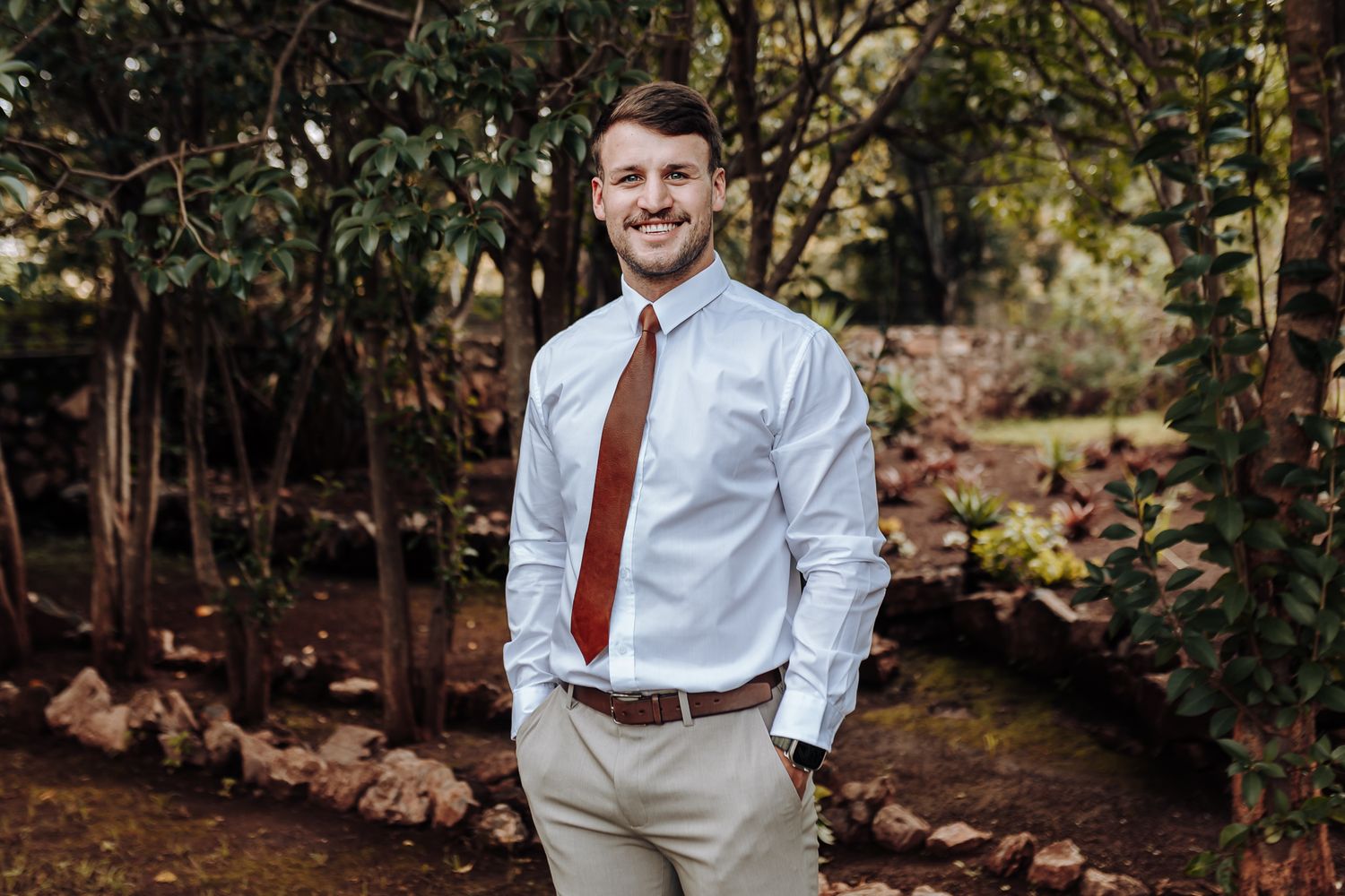 Business casual portrait featuring man in white shirt and brown tie against forest background.