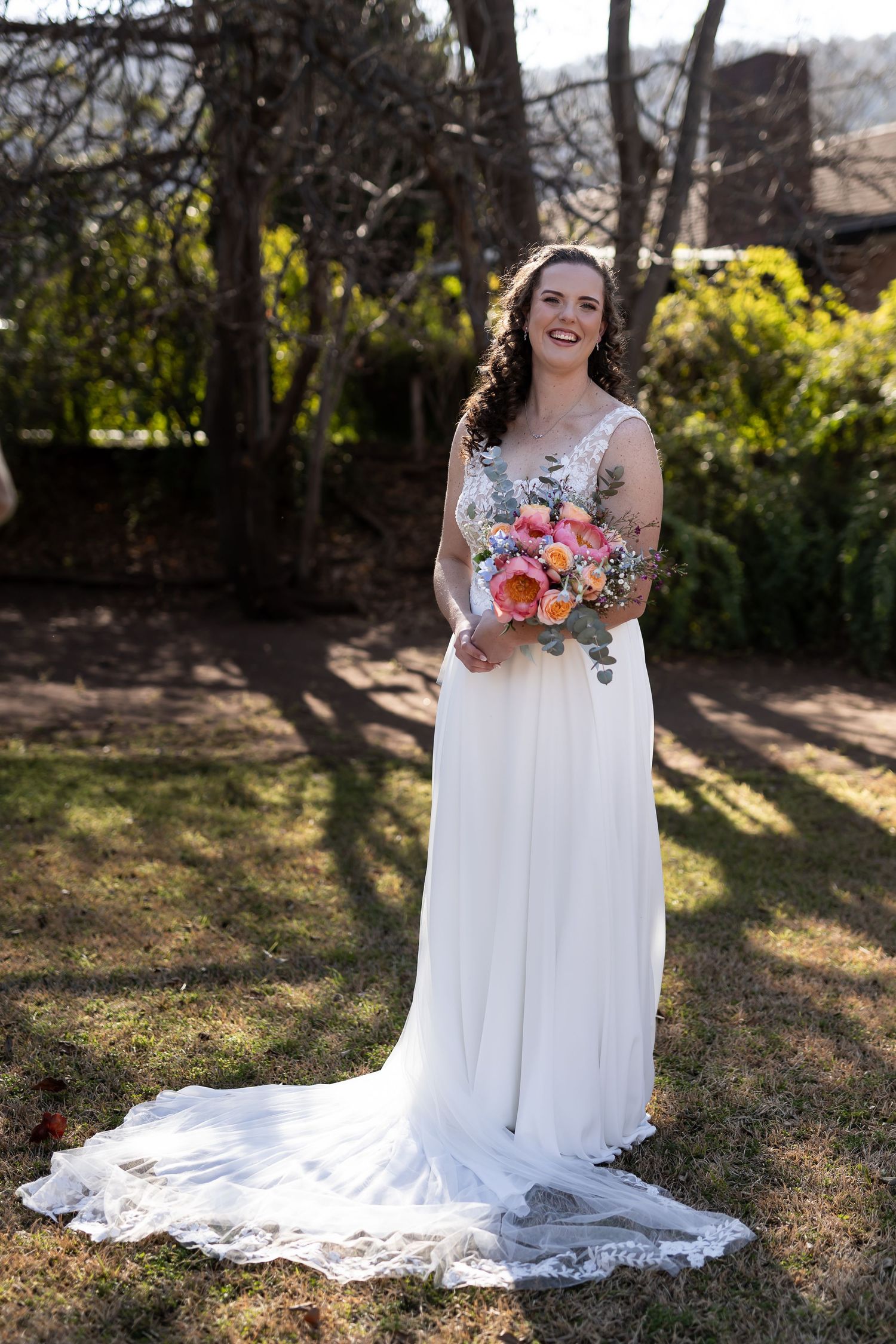 Bride in flowing white wedding dress holding vibrant coral and orange bouquet poses in sunlit garden setting.