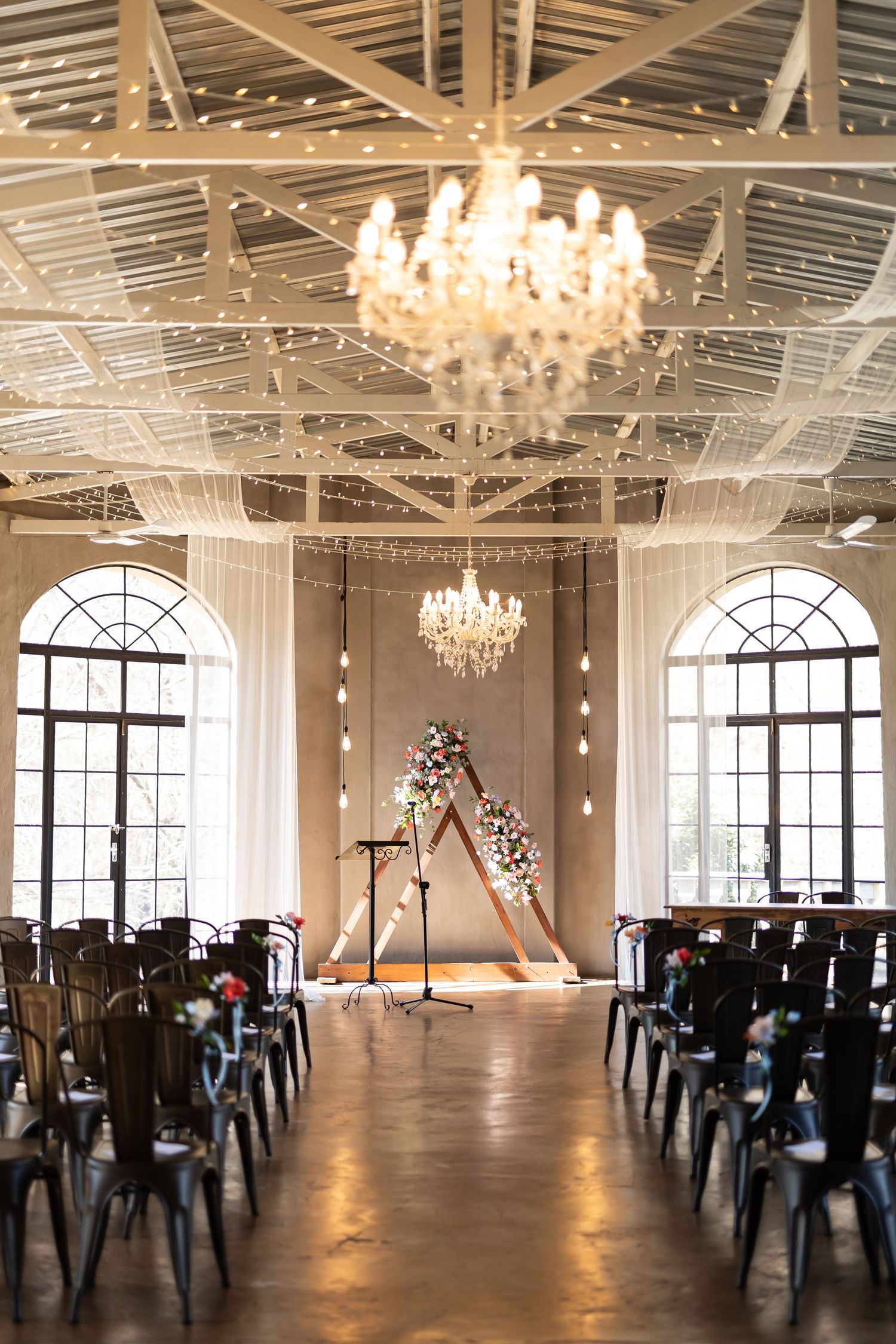 Elegant wedding ceremony setup in industrial venue with crystal chandeliers, arched windows and floral triangle arch.