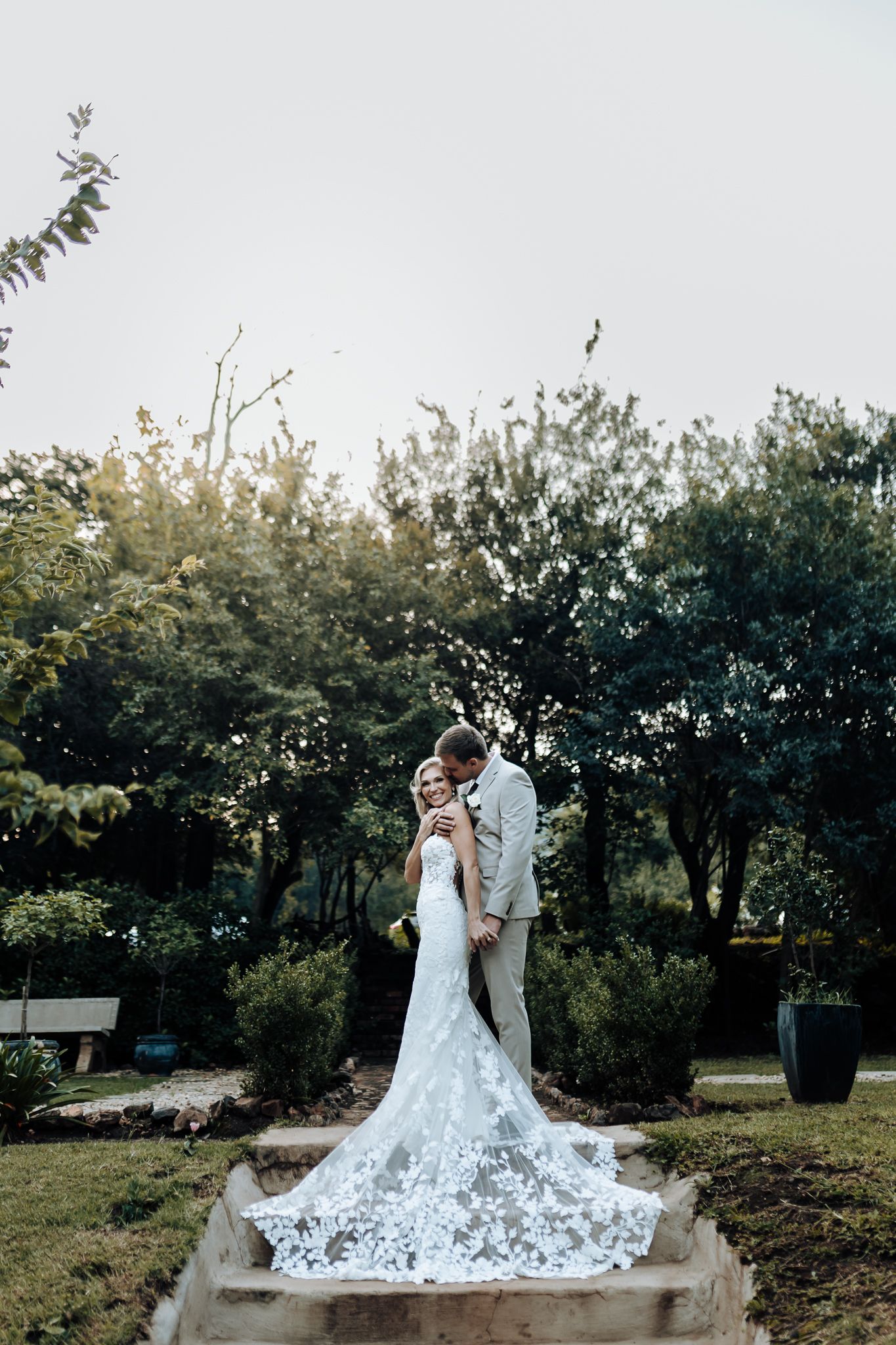 Wedding couple embracing on stone path with dramatic lace train flowing behind.