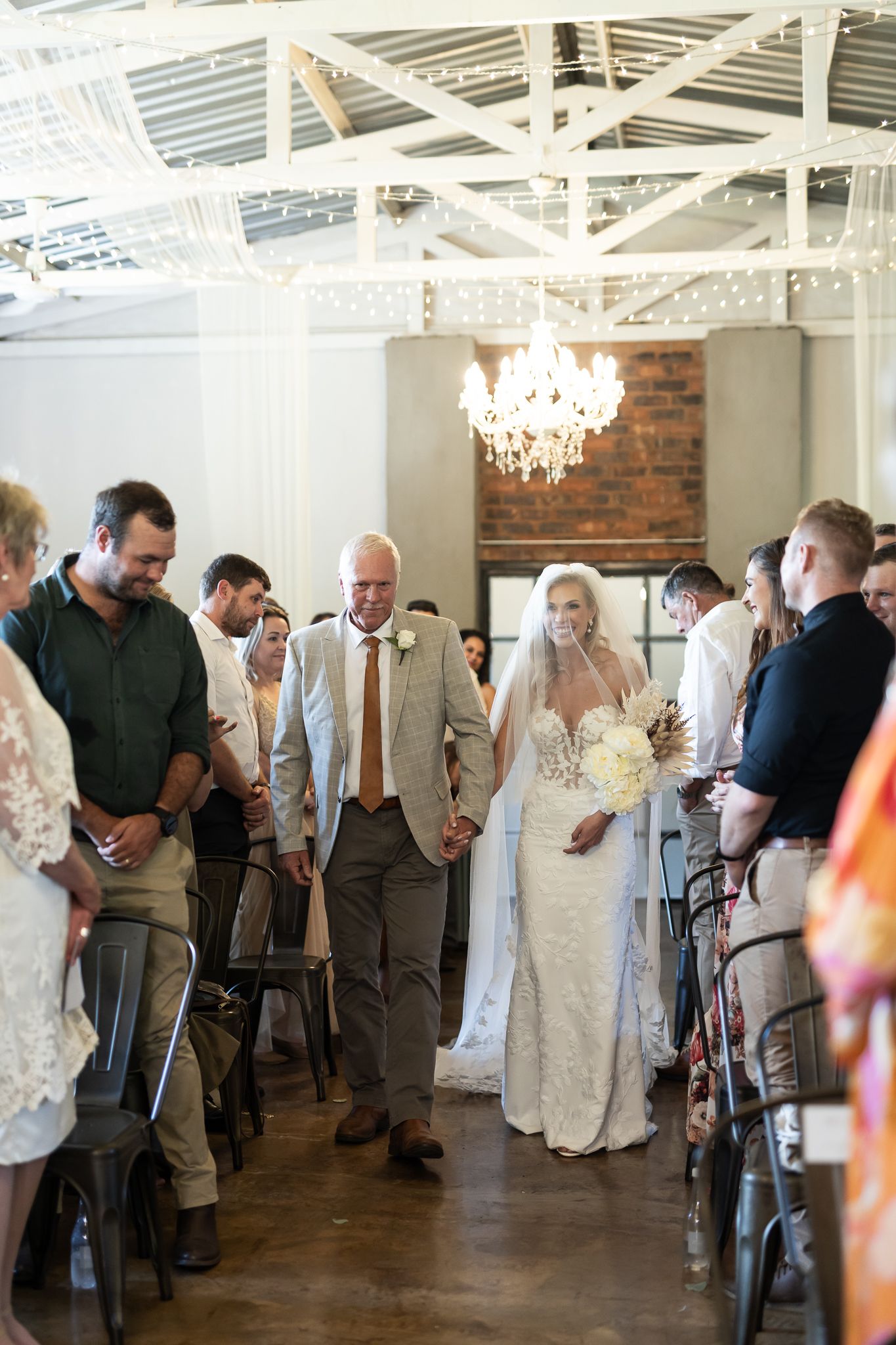 Wedding party in formal attire stands at front of ceremony venue with arched windows.