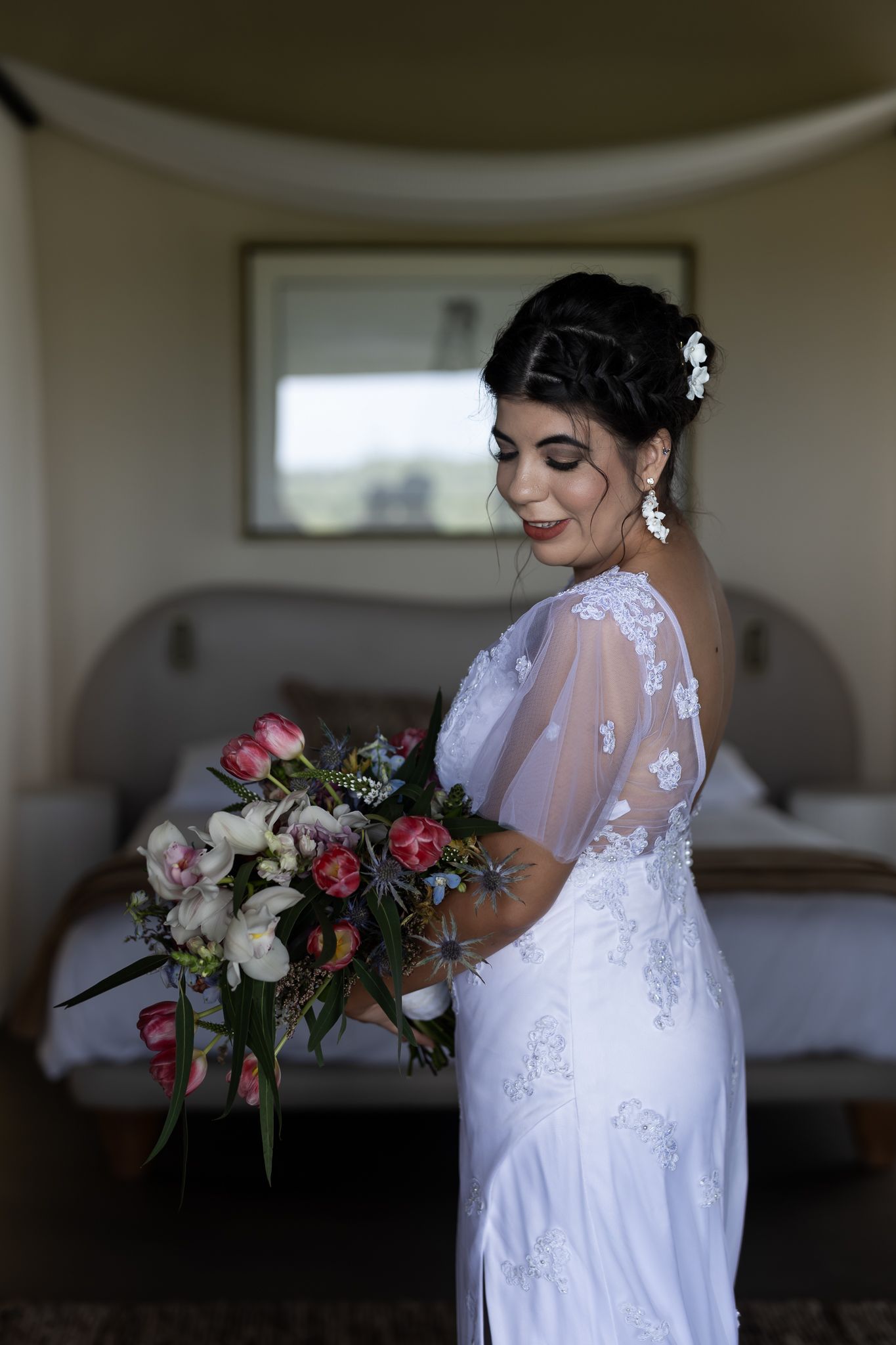 A bride in a white lace dress with sheer sleeves holds a pink and white bouquet while standing in a bedroom.