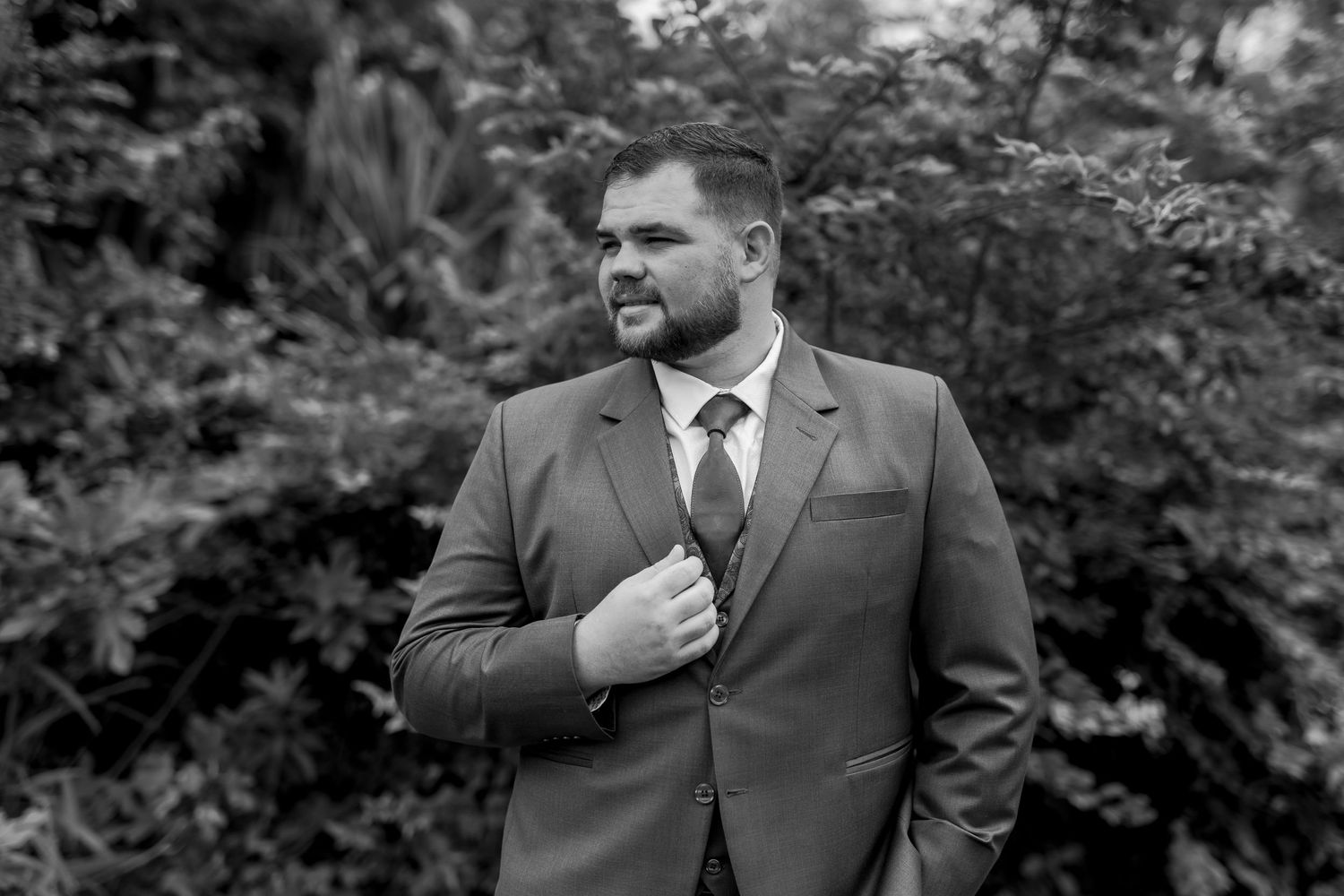Black and white portrait of a groom in a suit adjusting his tie outdoors.