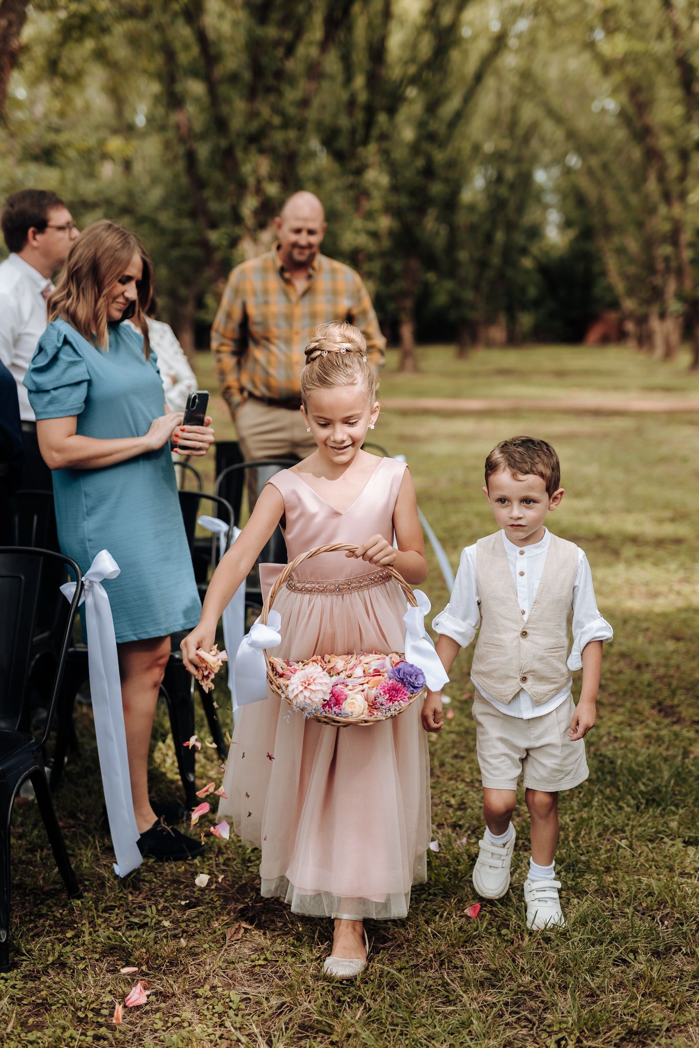 Young flower girl in pink dress and ring bearer in white walk down garden path during wedding ceremony.