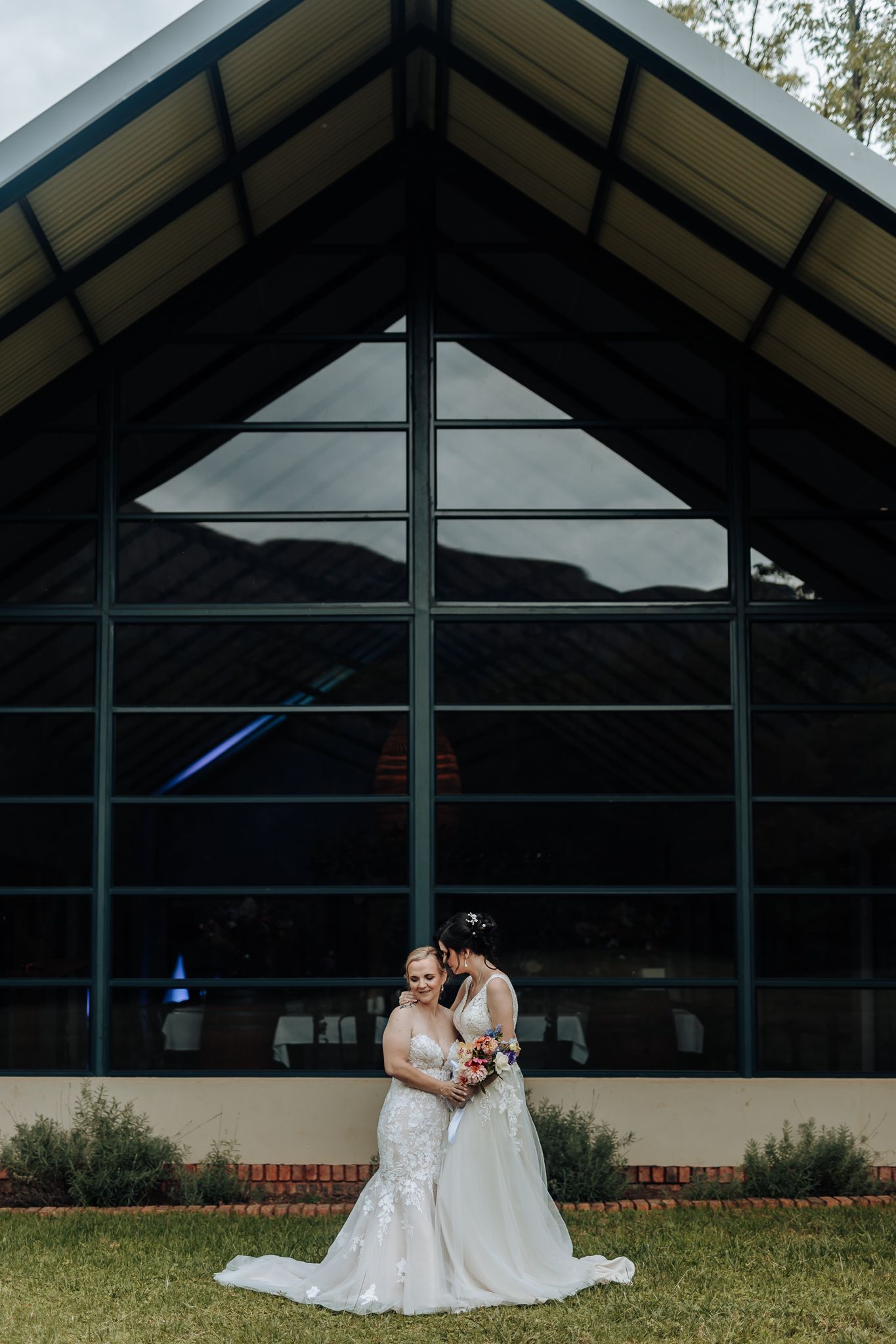 Two brides embrace outside modern chapel with dramatic angular architecture and mountain backdrop.