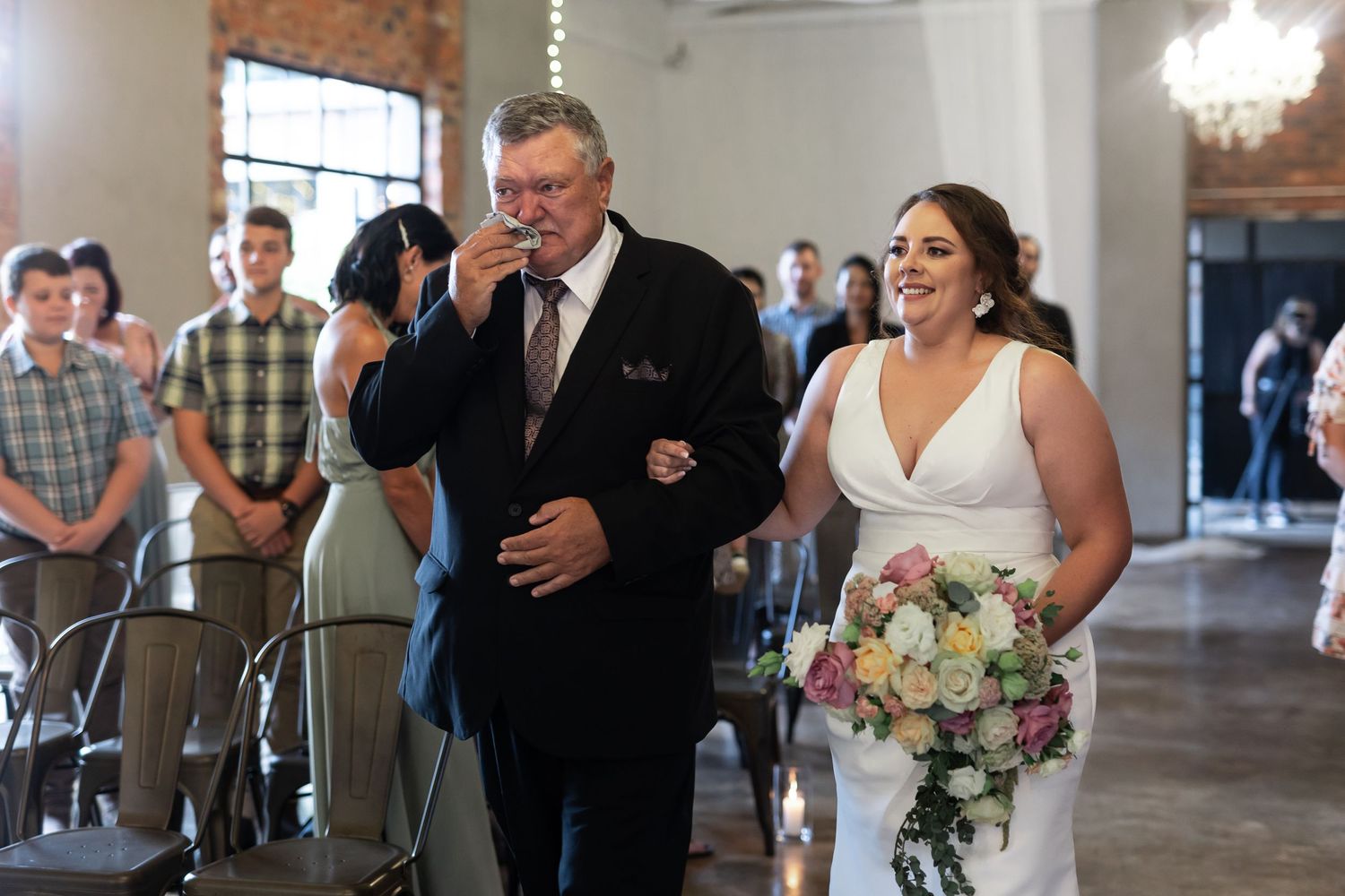 A bride in a white dress holds a pastel bouquet while walking down the aisle during an indoor ceremony.