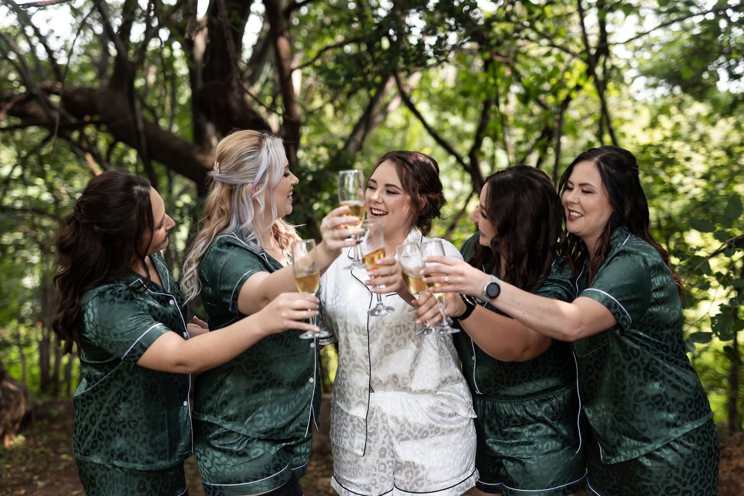 Wedding party in coordinated outfits toasting with champagne glasses in a forest setting.