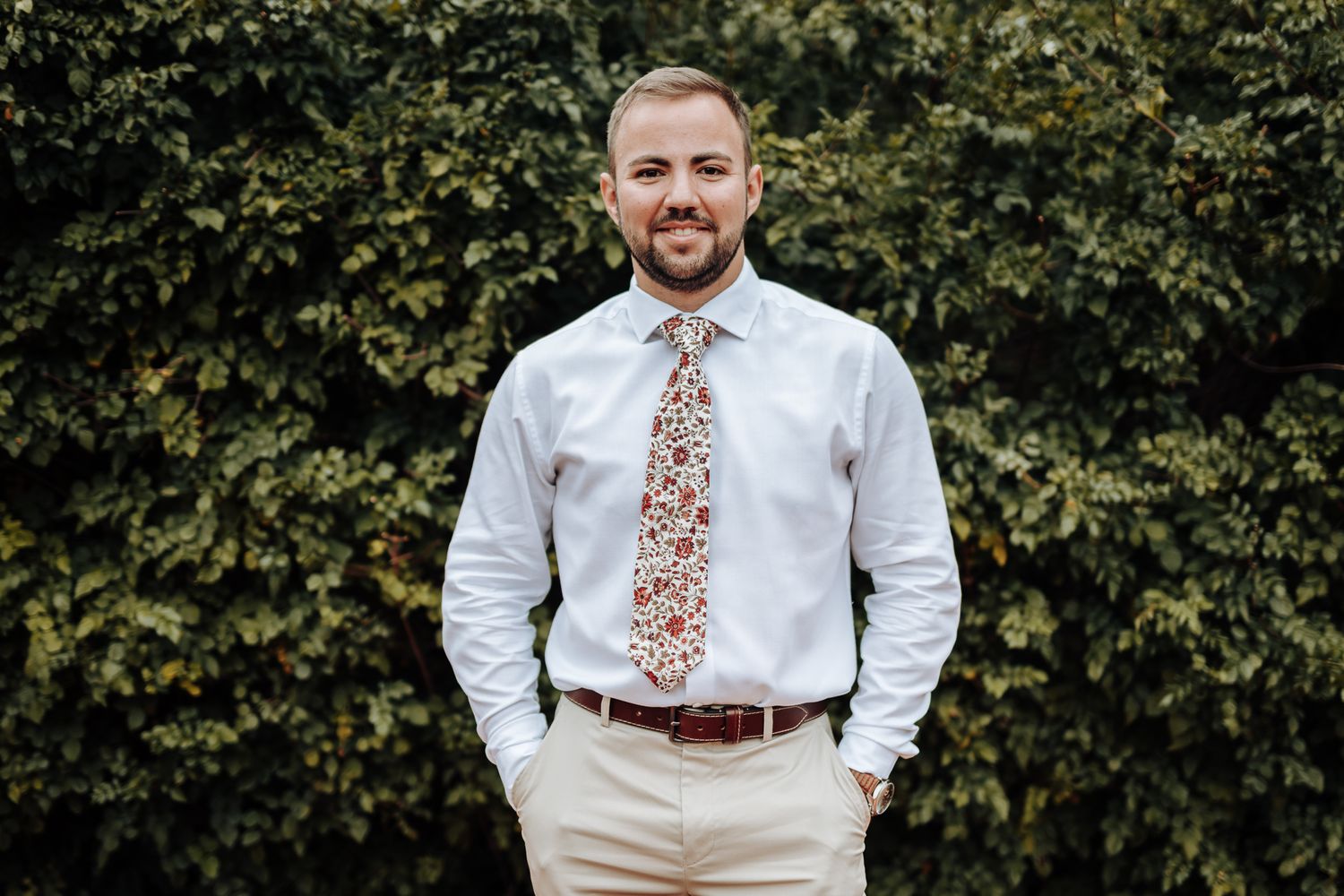 Groomsman wearing white shirt and floral tie poses in front of green hedge.