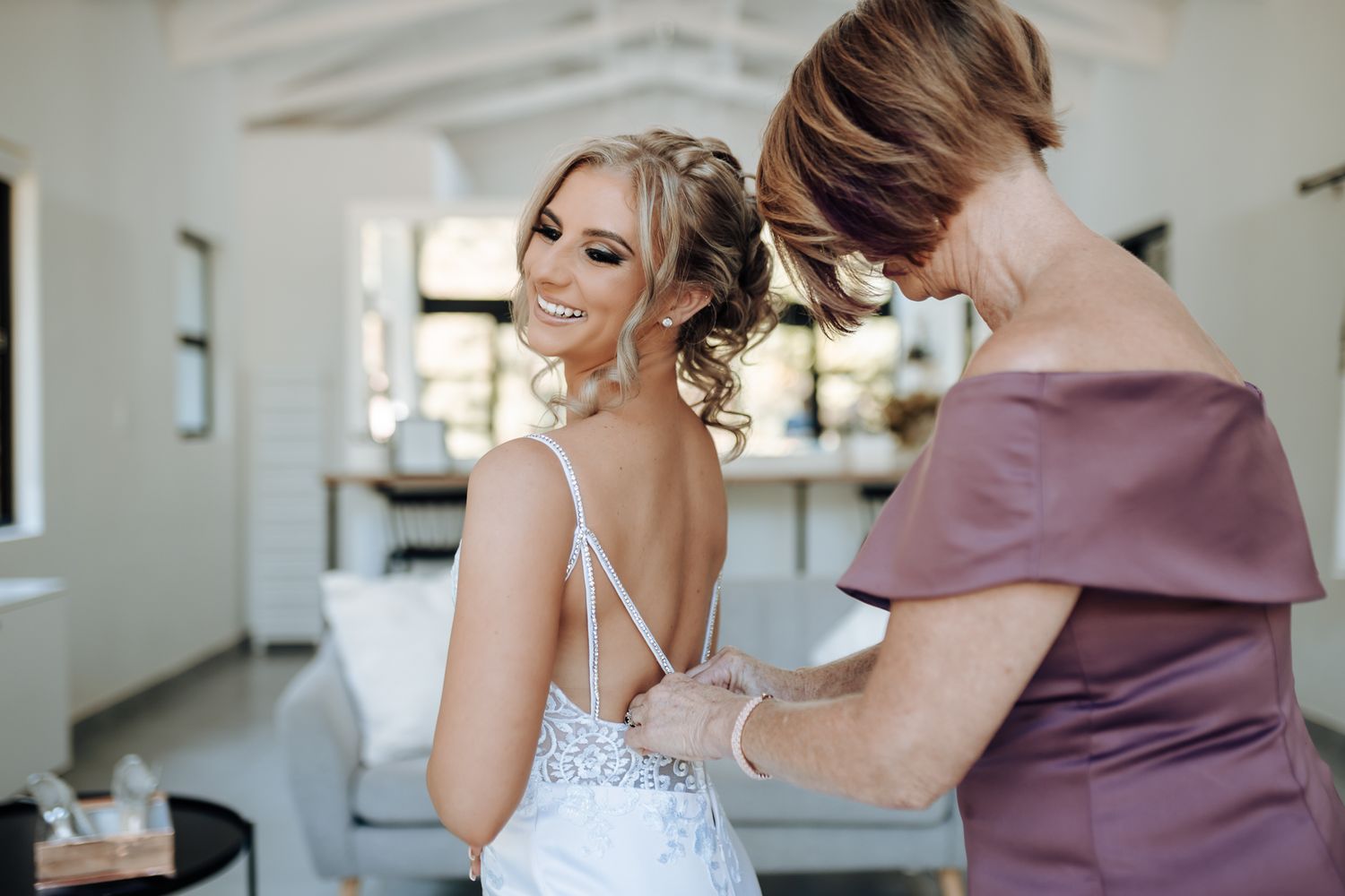Person assists with final adjustments to wedding dress in elegant white room with exposed beams.