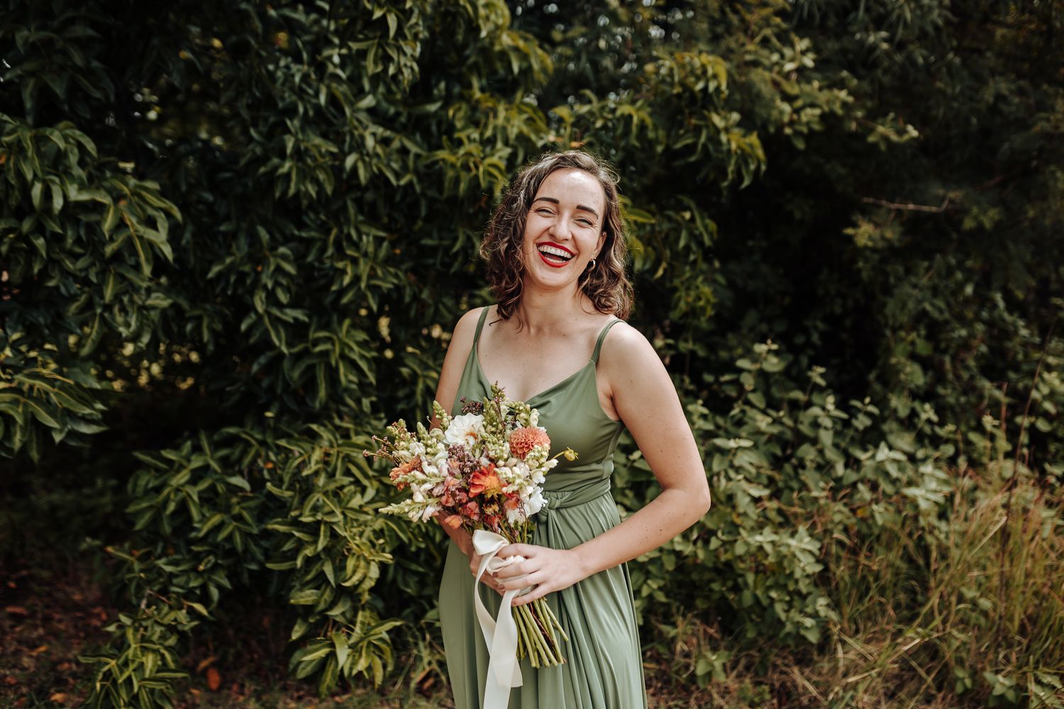 Person in sage green dress holding coral and white bouquet while laughing outdoors.