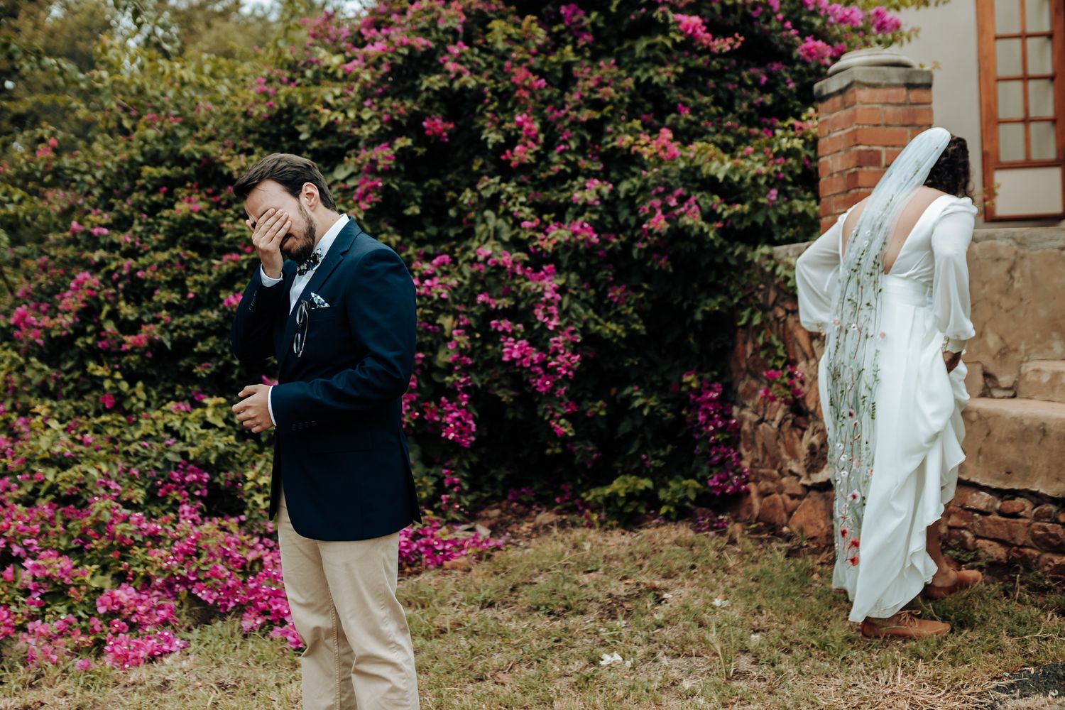 First look moment captured with couple standing separately against pink flowering bushes.