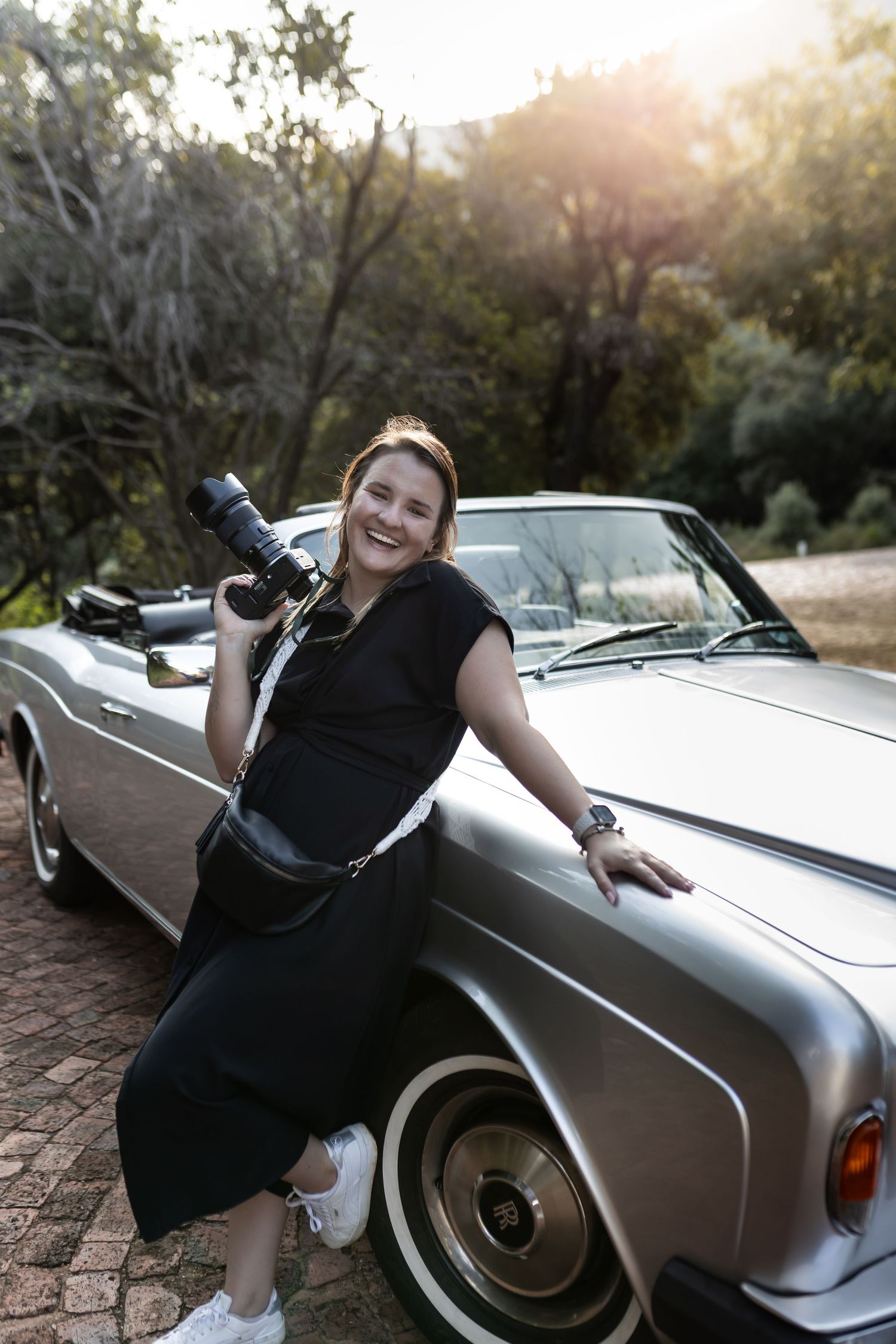 Professional photographer in black dress poses with camera beside vintage silver convertible.