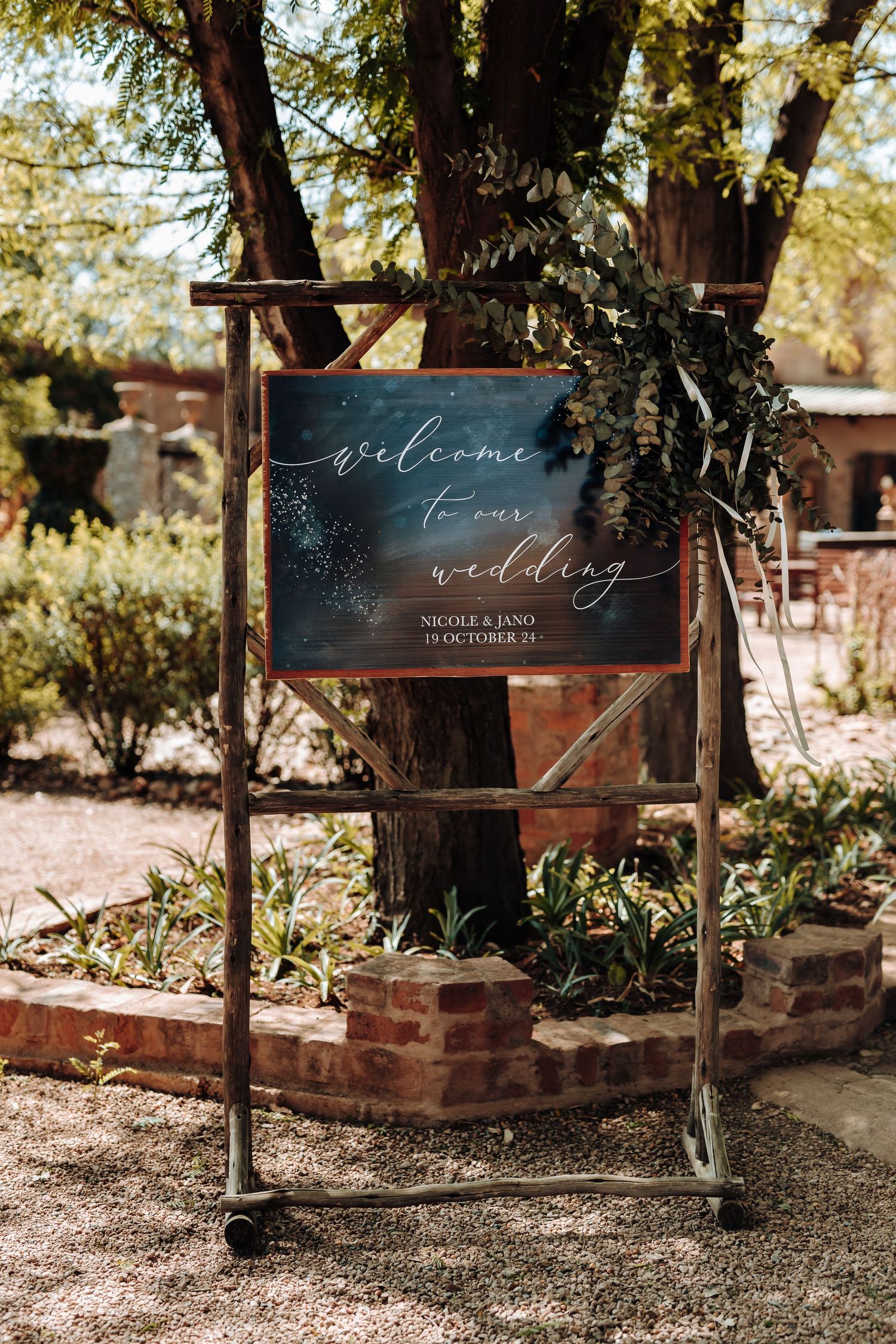Rustic welcome wedding sign displayed on wooden stand against garden backdrop.