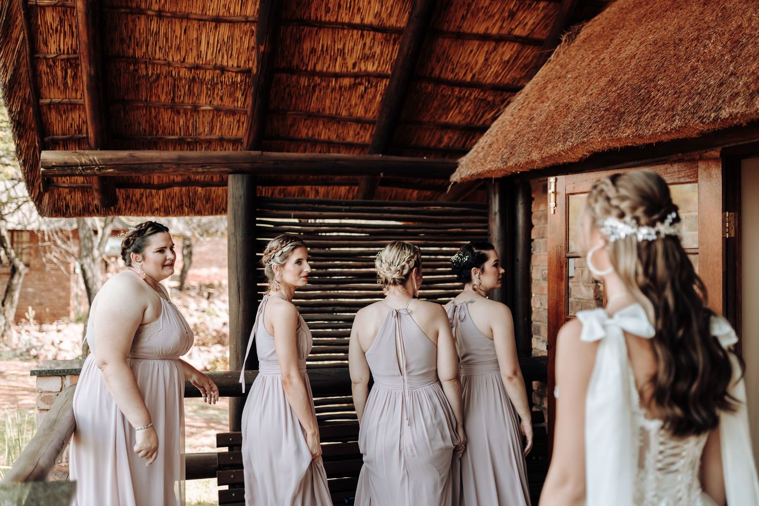 Wedding party poses under a thatched roof with bridesmaids in blush gowns and bride in white.