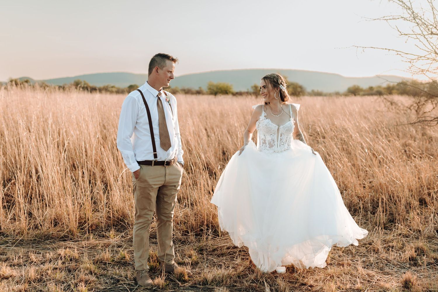 Wedding couple poses playfully in rustic field with mountains and setting sun.