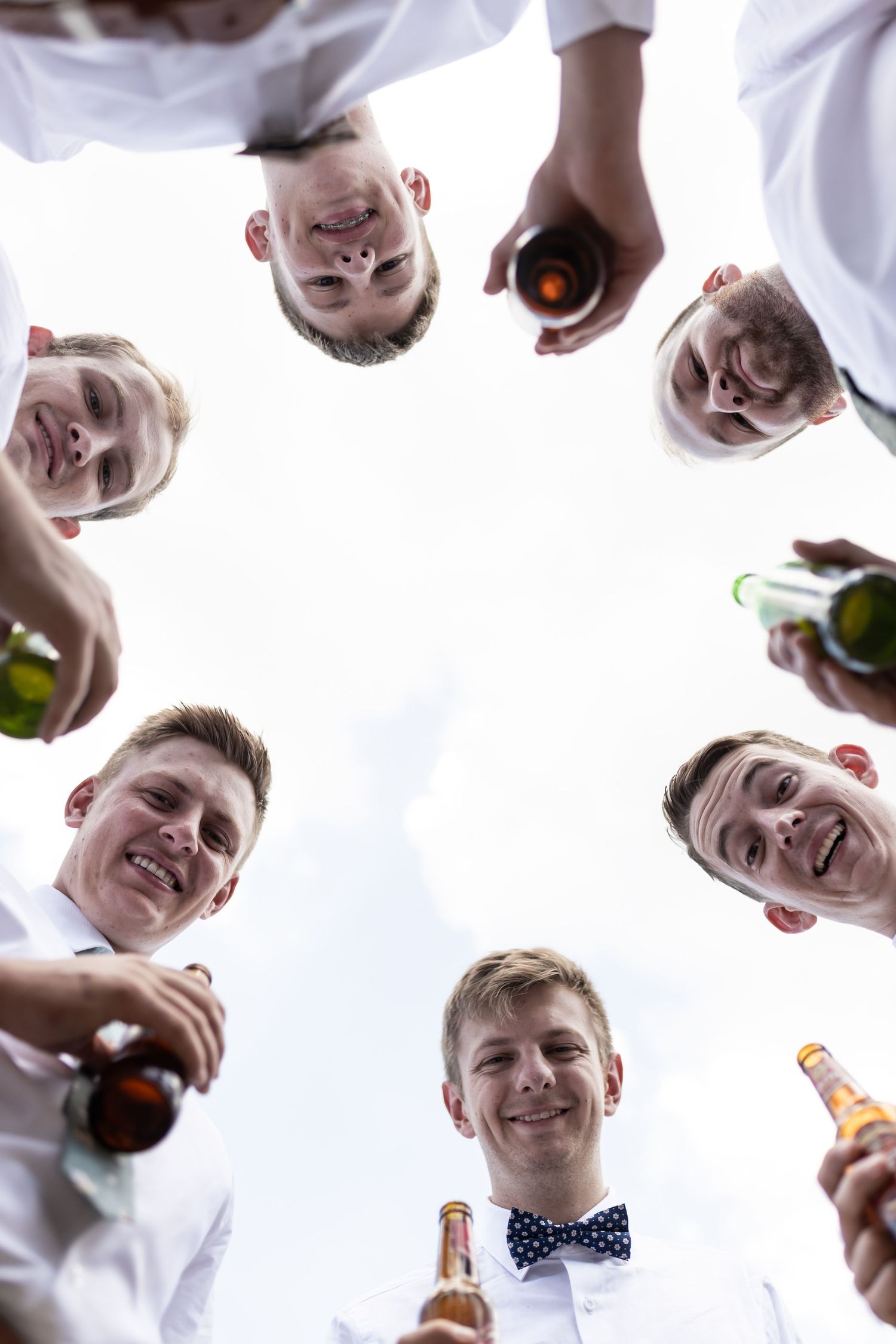 Circle view from below of groomsmen toasting with bottles against bright sky.