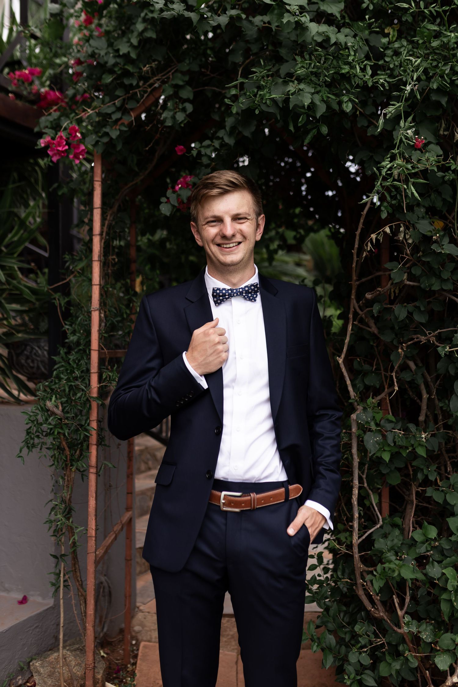A formal portrait in front of blooming bougainvillea showing a dapper bow tie and navy suit combination.