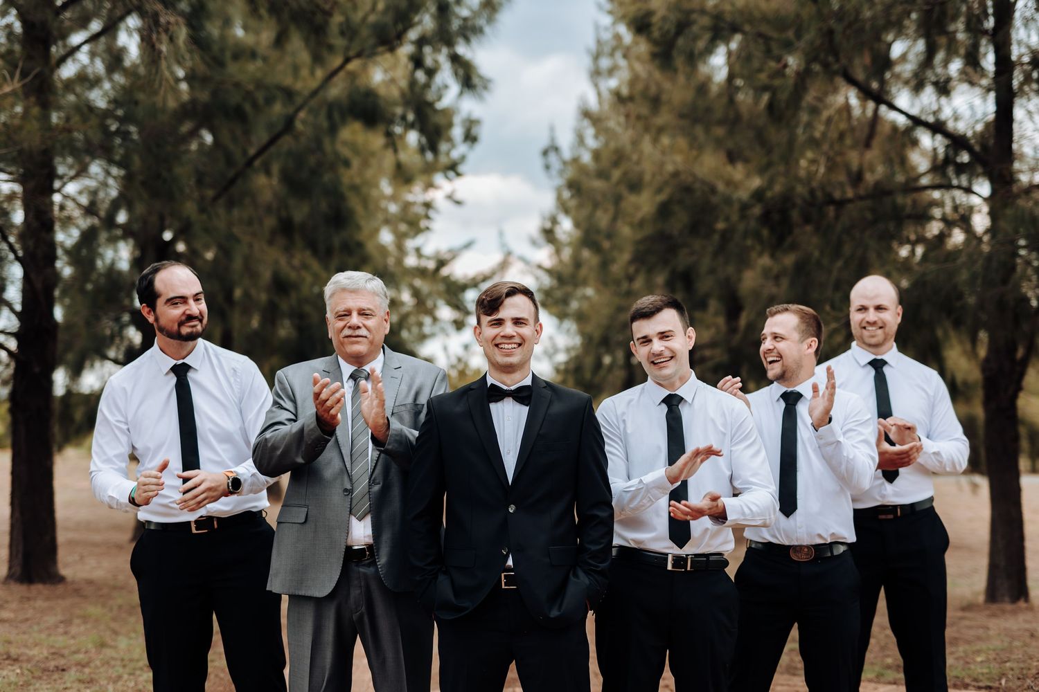 Wedding party members gesture and pose casually while standing among pine trees.