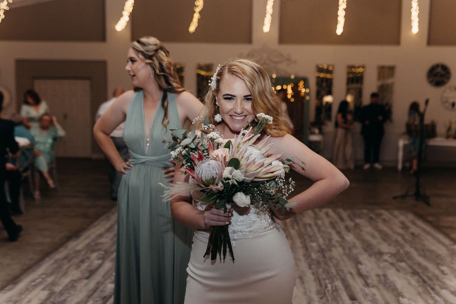 A woman in a white dress holds a beautiful bouquet while another in sage green stands nearby.