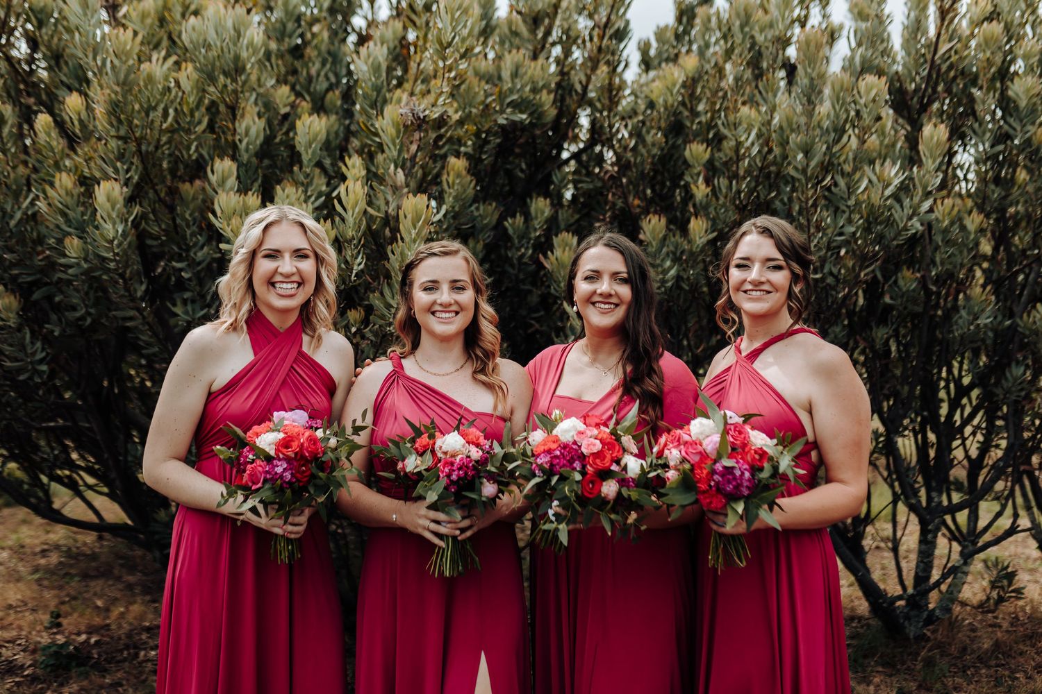 Four bridesmaids in matching red dresses pose with floral bouquets against evergreen backdrop.