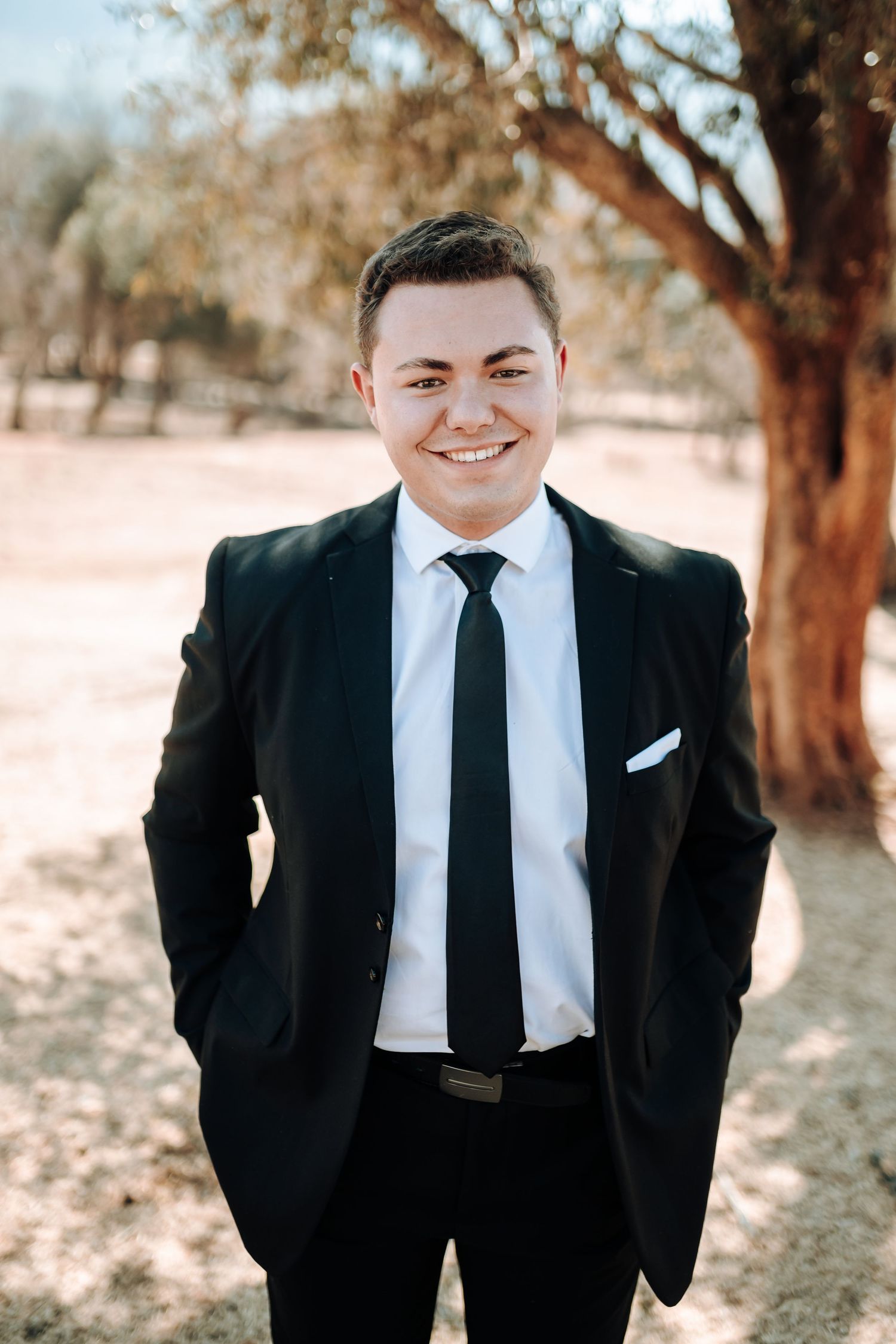 Professional portrait of someone in a black suit against a natural outdoor background.