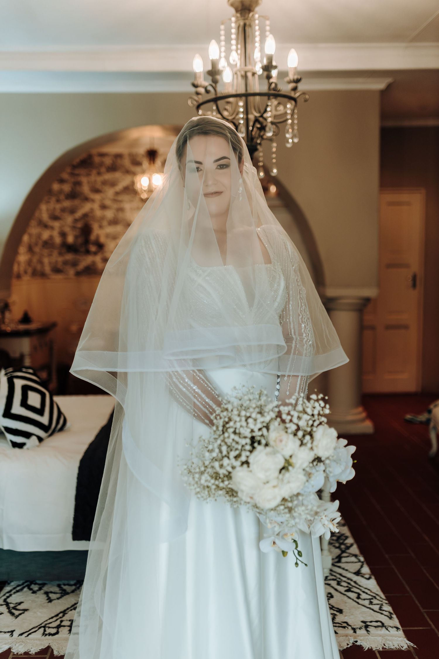 Dreamy bridal photo with layered veil and white floral bouquet under crystal chandelier lighting.