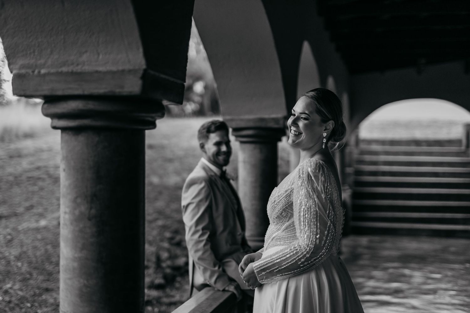 Black and white photo of newlyweds sharing a joyous moment under classic stone columns.