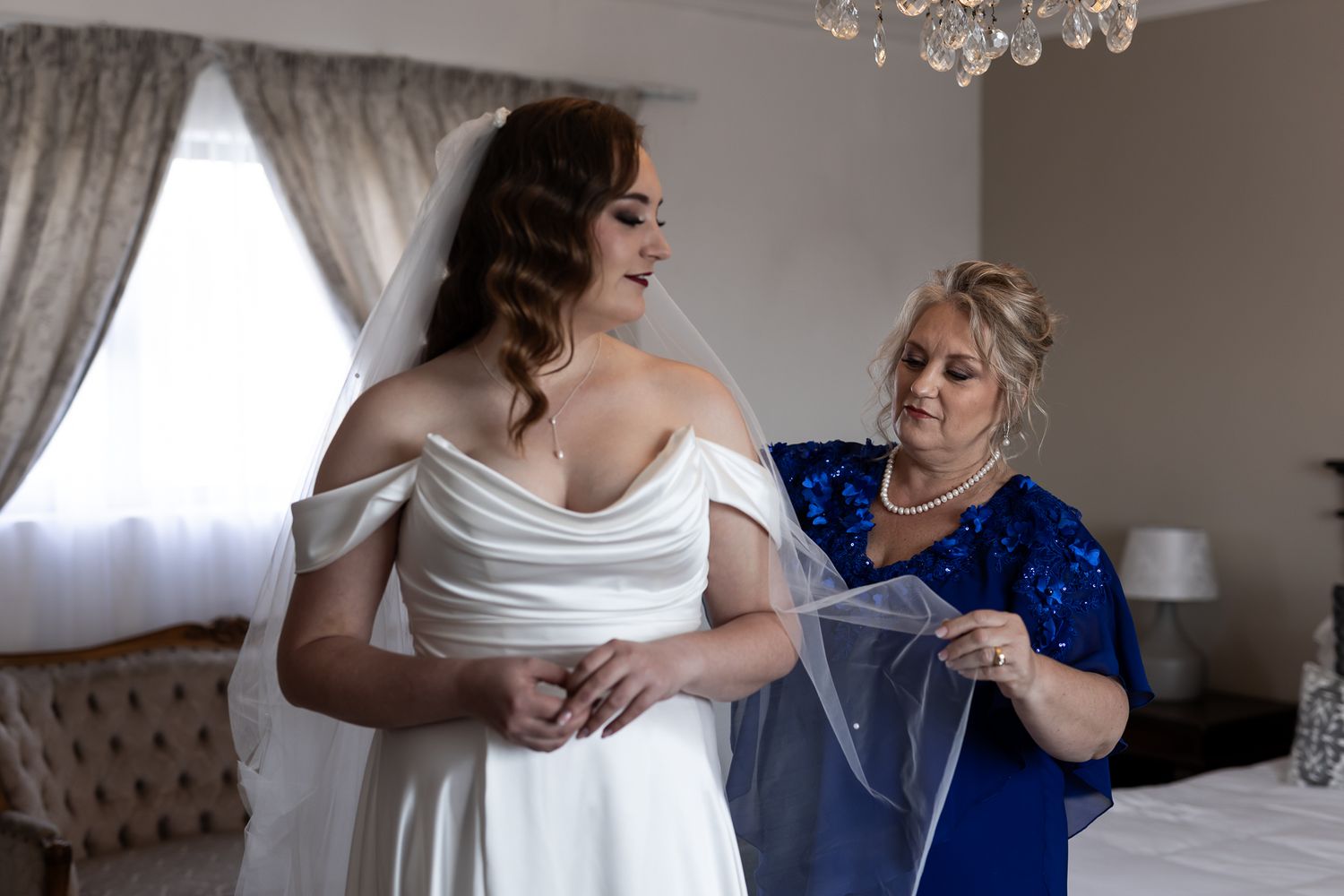 Wedding preparation scene with helper in royal blue dress adjusting bridal veil.