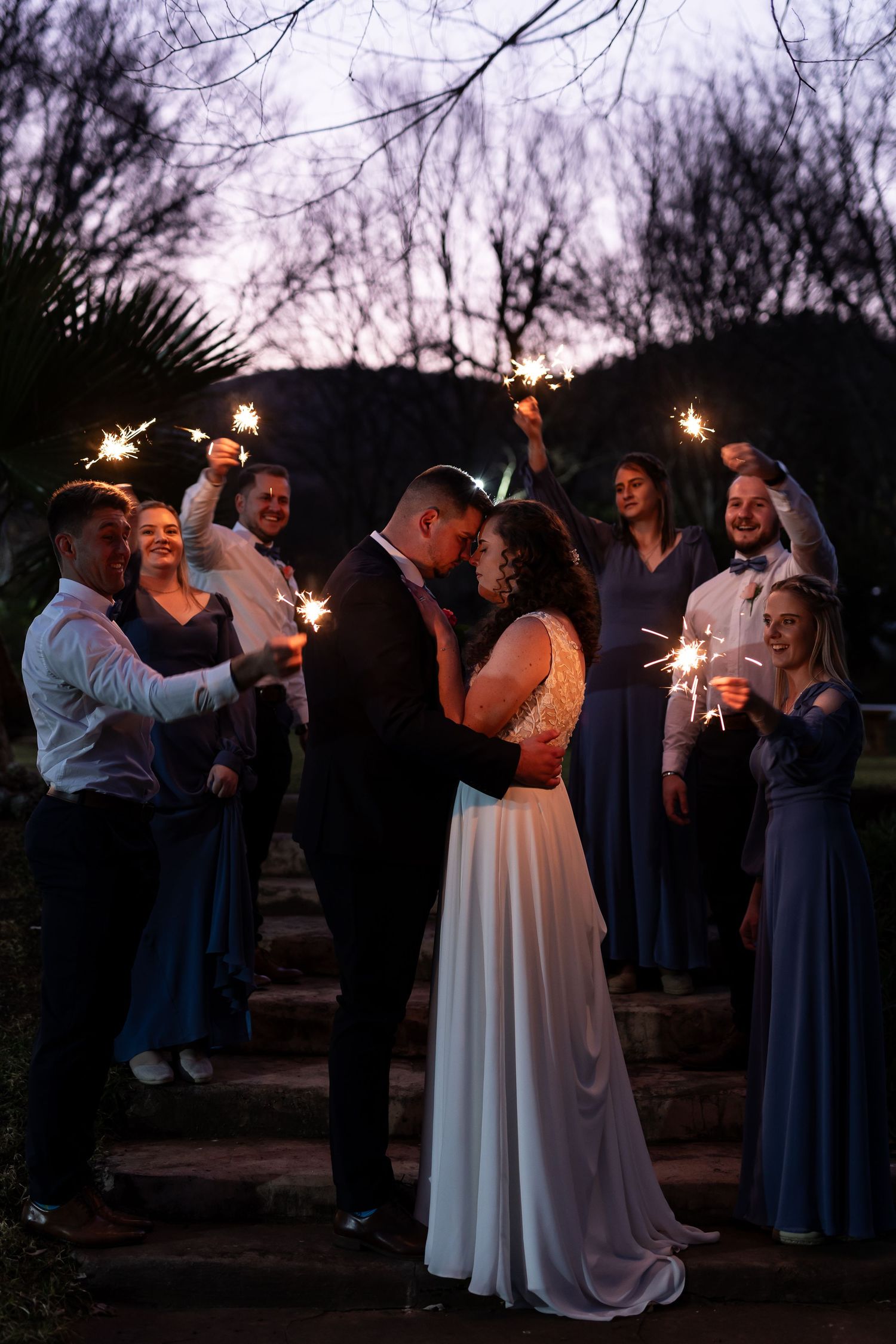A romantic wedding couple dances at dusk surrounded by guests in navy blue attire during an outdoor reception.