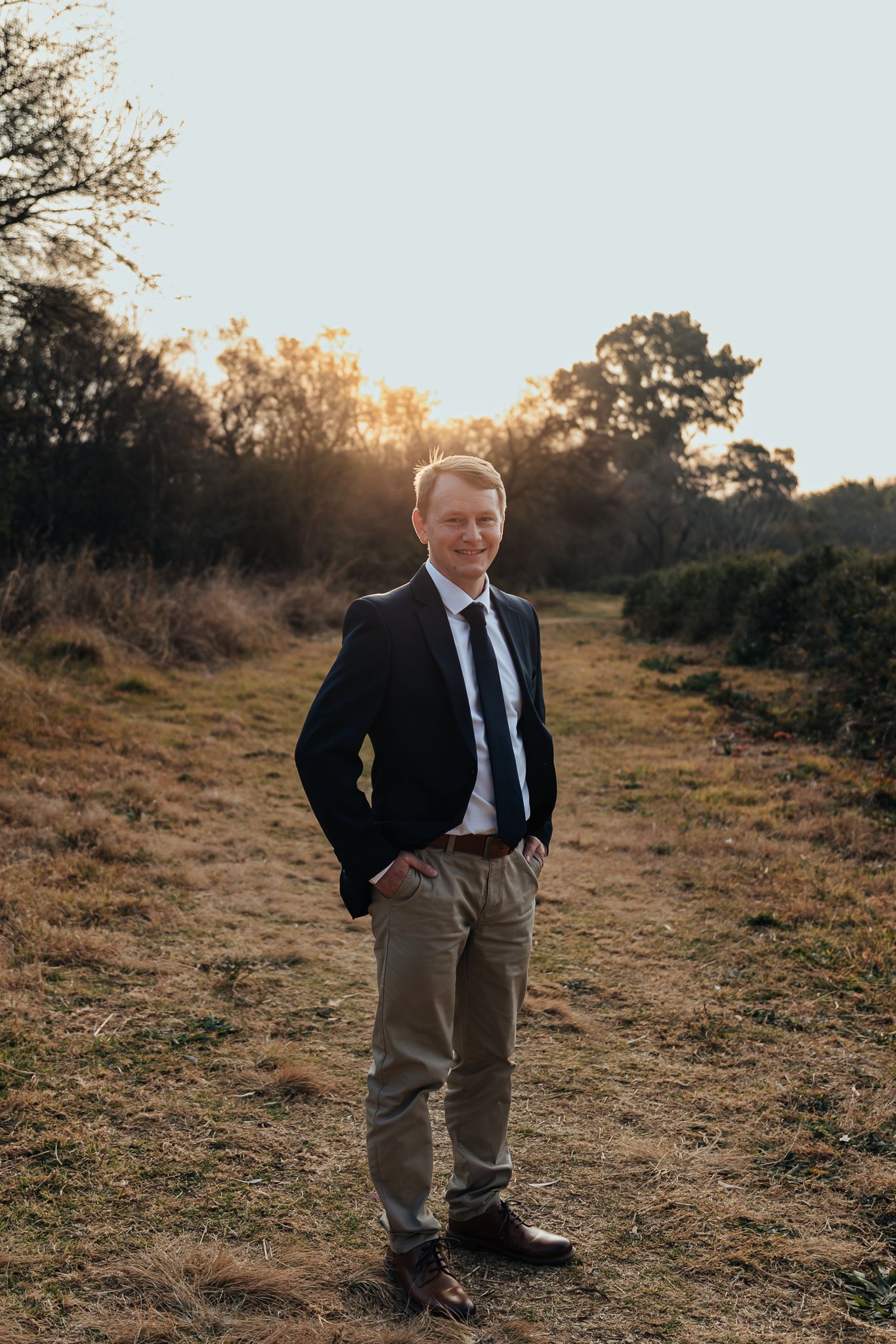 A groom in a dark suit and khaki pants poses on a dirt path at sunset with trees in the background.