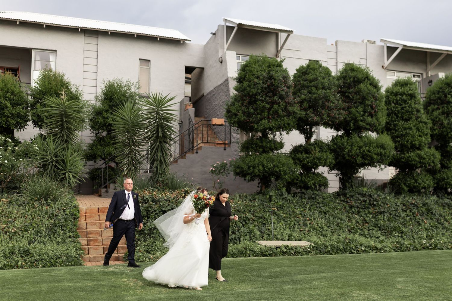 A wedding couple walking across a manicured lawn with modern architecture in the background.