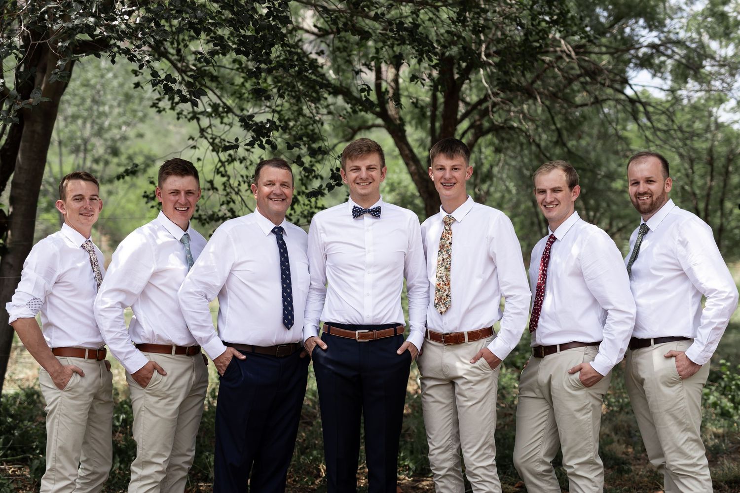 Wedding party in coordinated white shirts and khakis poses informally among trees.