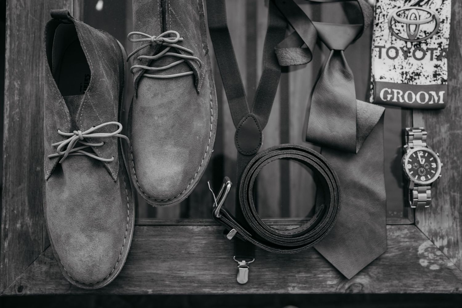 Classic men's accessories including brown suede desert boots, leather belt, tie and watch arranged on wooden surface.