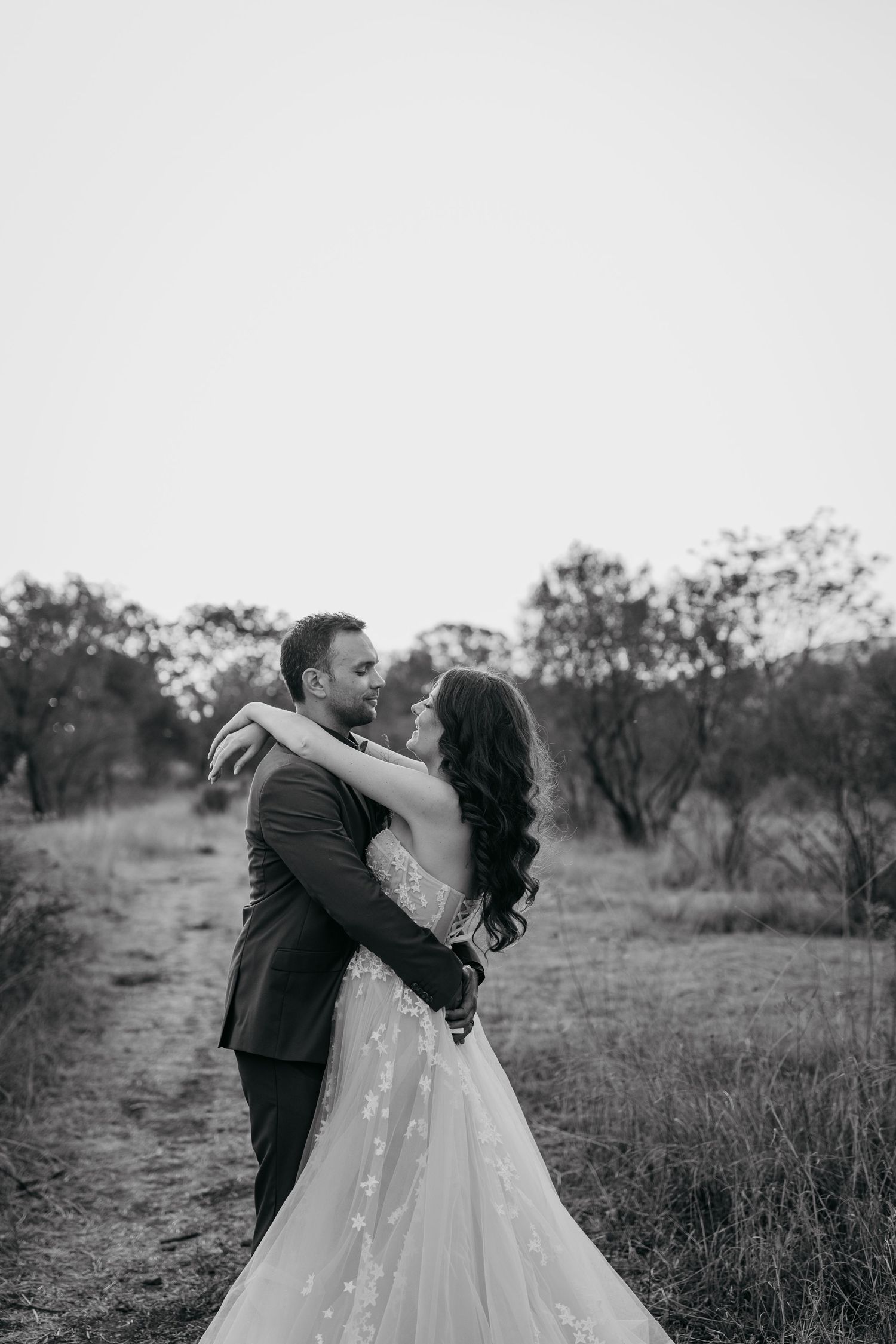Black and white portrait of a couple embracing on a rural path surrounded by trees.