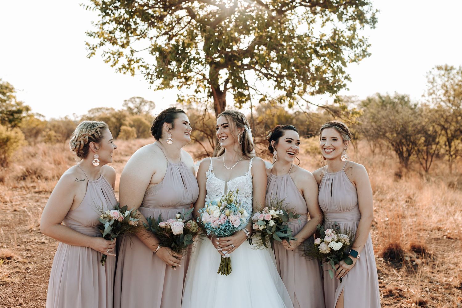 Bridal party laughing together in mauve dresses against golden sunset landscape.