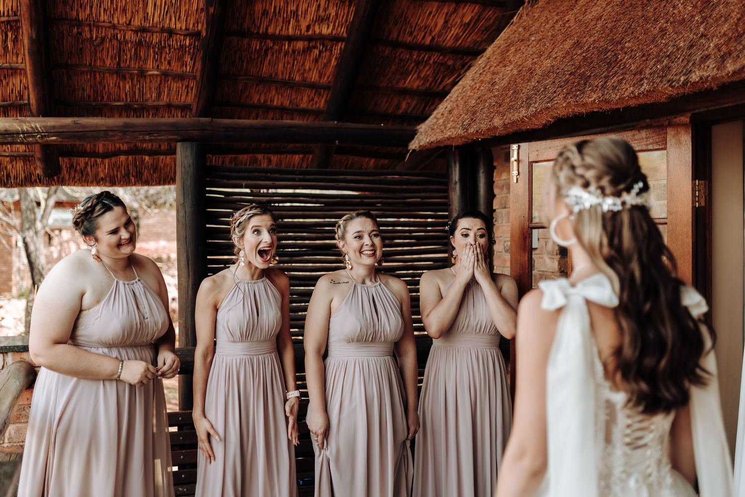 A bride in white reveals her dress to bridesmaids in matching blush gowns under a rustic thatched roof.