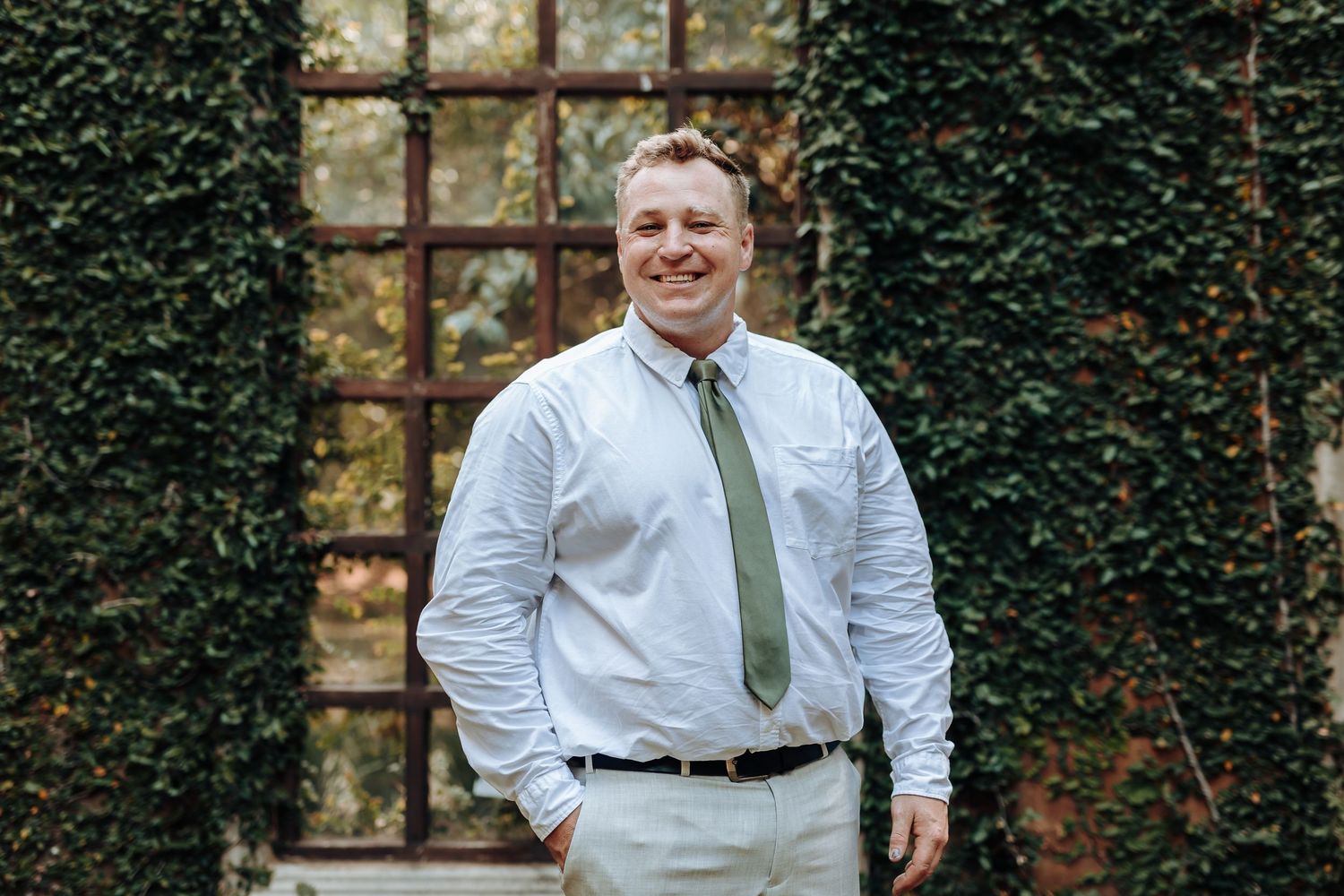 Groom in light blue shirt and sage green tie poses against ivy-covered wall with vintage window frame.