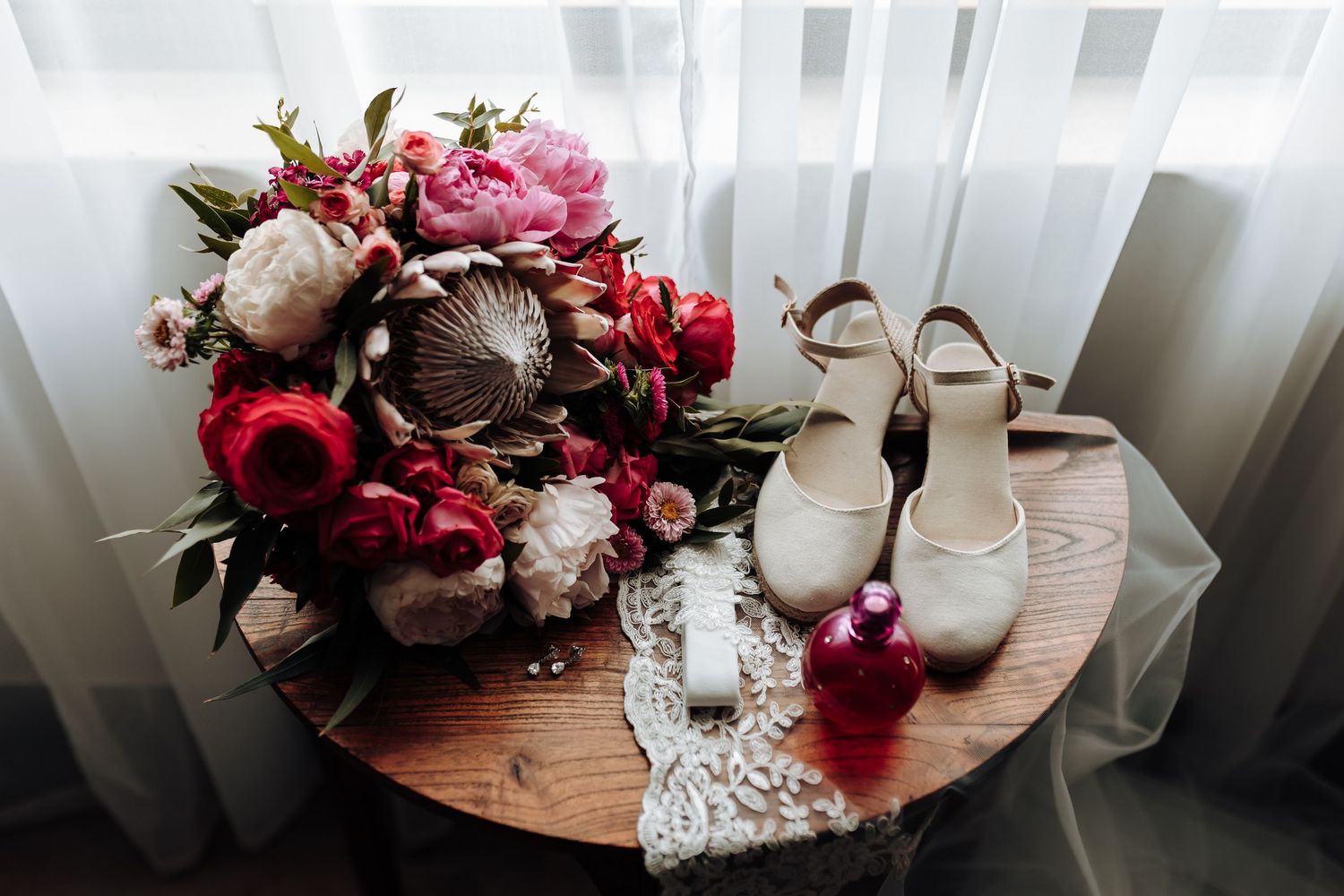 Wedding details featuring pink bouquet, white shoes and lace accessories on wooden table.