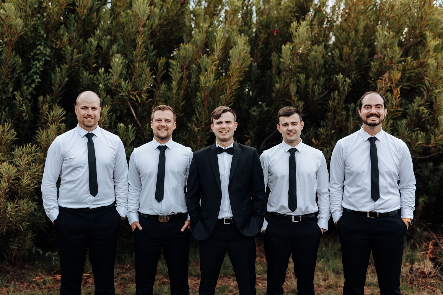 Wedding party in formal attire poses together against an evergreen backdrop.