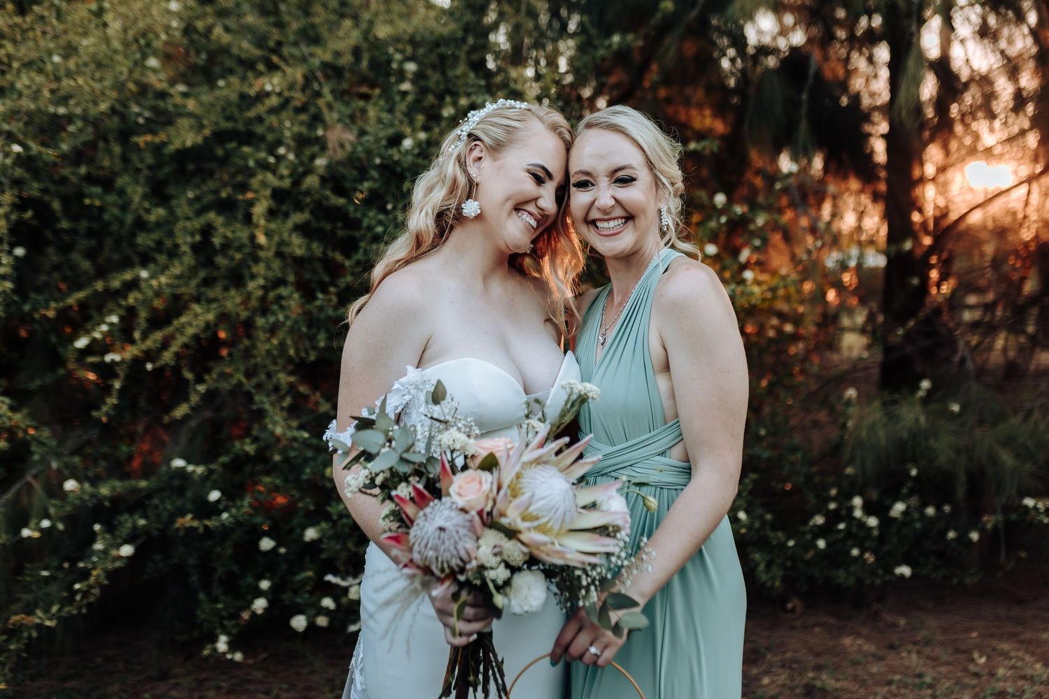 Bride in white dress and bridesmaid in mint green gown share joyful moment with beautiful floral bouquets at sunset.
